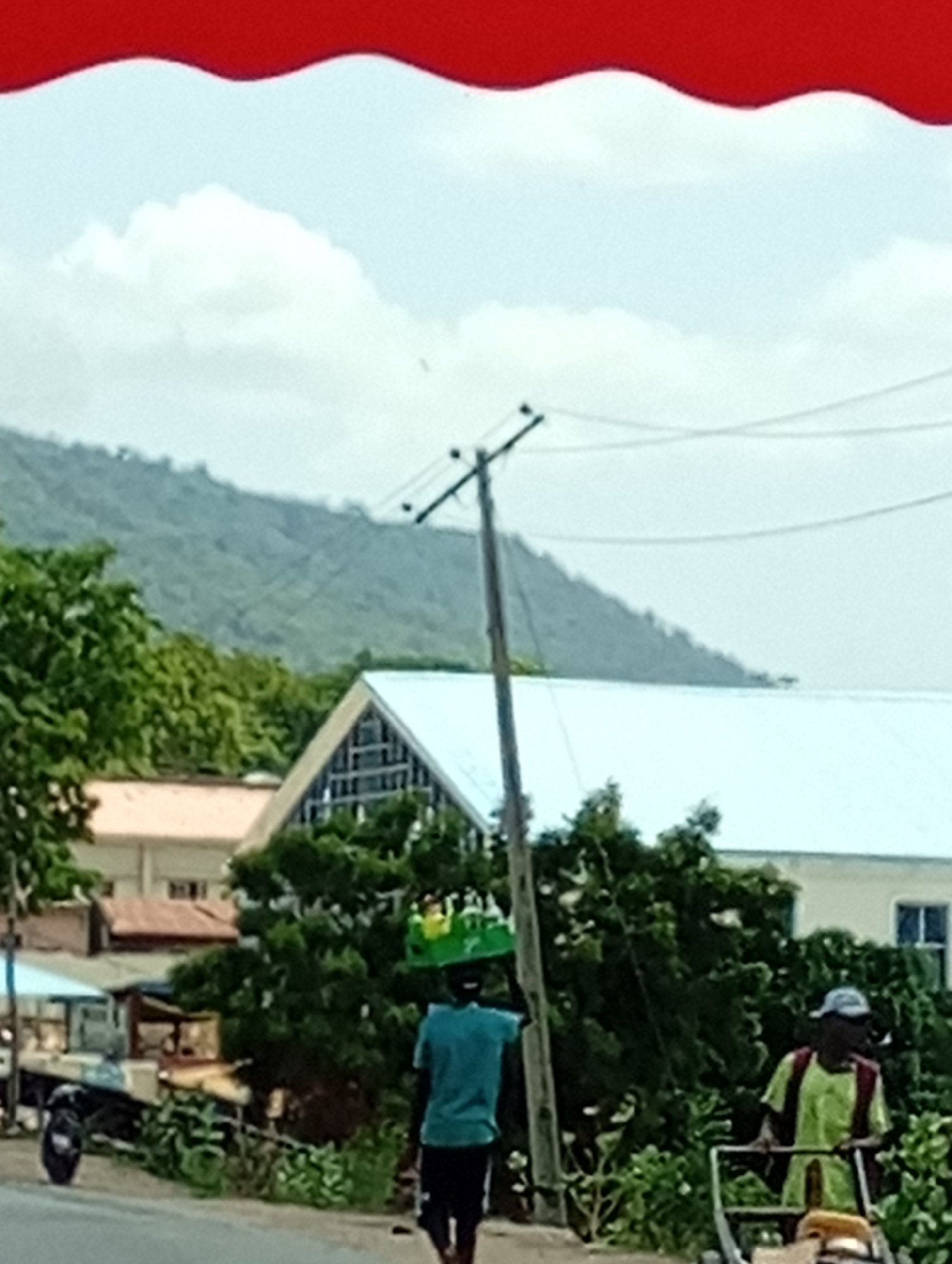 The image shows an outdoor scene with a man carrying what appears to be bottles or containers on his head, walking on a road or street. Another person is visible in the background, using what looks like a pushcart or gardening tool. There are buildings, trees, and an electrical pole in the background. A mountain range can be seen in the distance under a cloudy sky. A red awning or overhang is partially visible at the top of the image. The location is Unknown, Unknown.