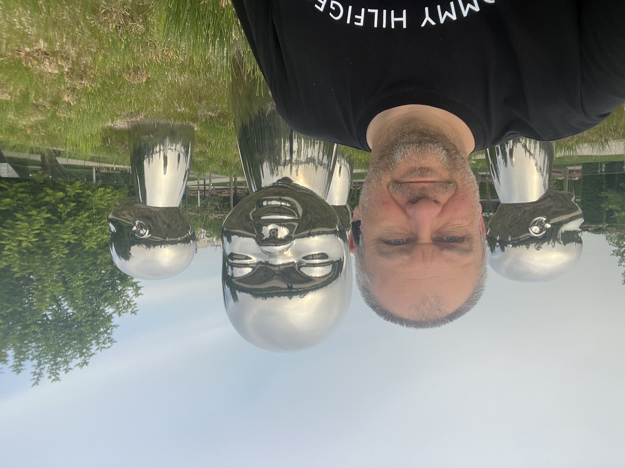The image shows a man standing in front of a public art installation featuring large, polished metal head sculptures in Krasnodar, Russia. The man is wearing a black Tommy Hilfiger t-shirt. The sculptures are reflective, mirroring the sky and surrounding environment. The background includes green grass and trees.