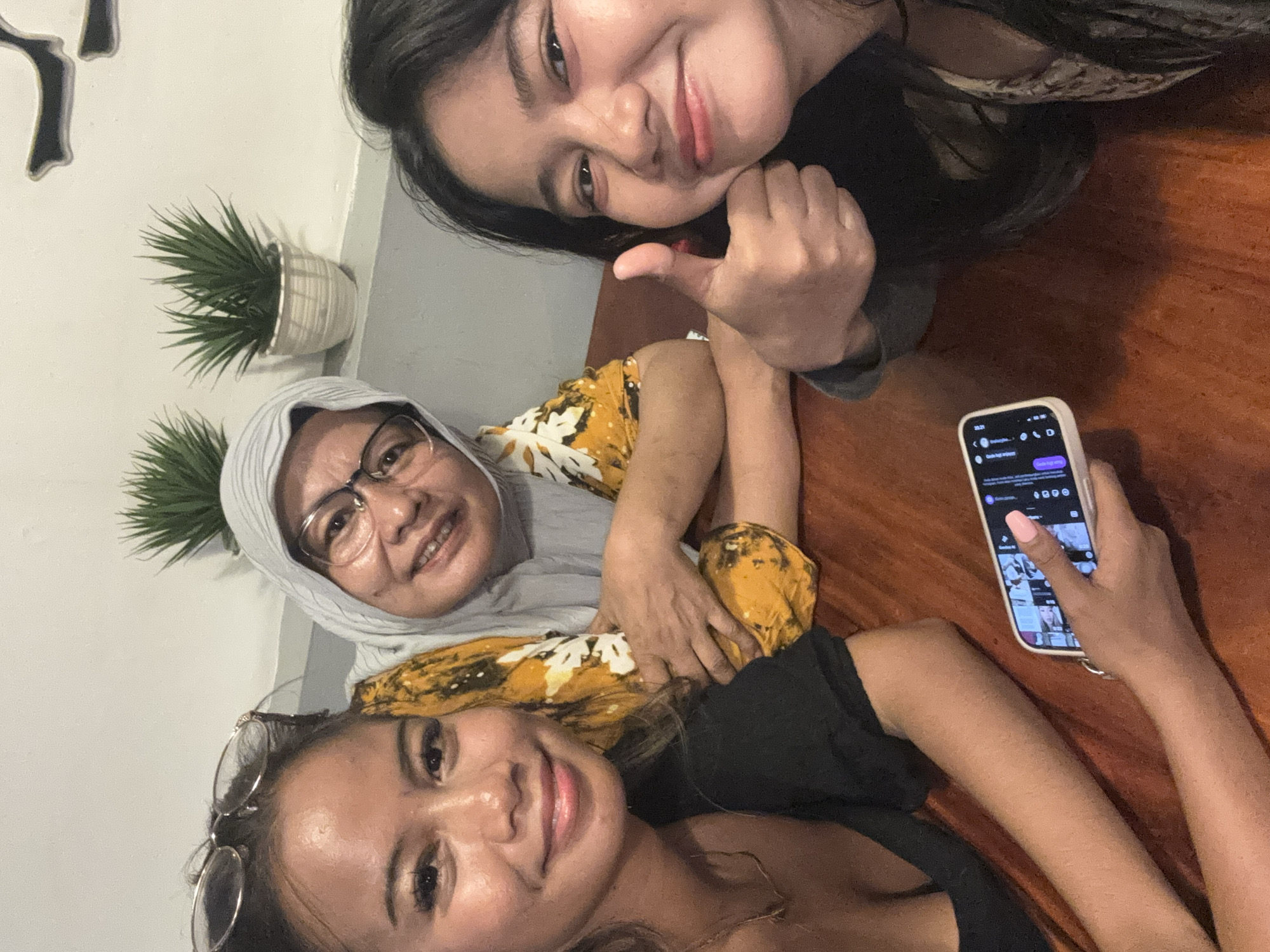 Three females are posing for a selfie in an indoor environment, with a wooden table as a prominent feature. The woman on the top right is giving a thumbs-up. A woman in a headscarf is in the middle. The third woman is on the bottom left and seems to be holding a mobile phone and scrolling. A potted plant is visible on the wall. The location is possibly North Jakarta, Indonesia.