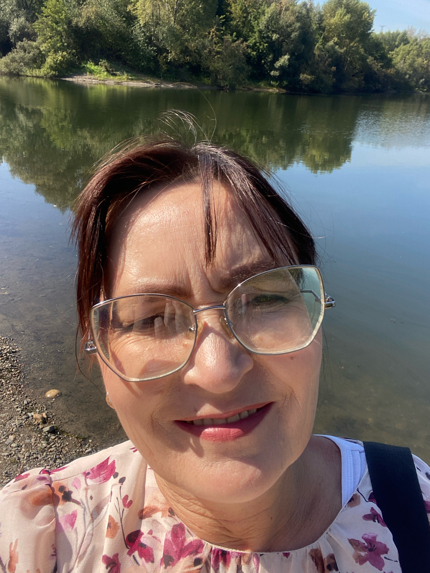 A woman with brown hair, wearing glasses and a floral print blouse, takes a selfie by a body of water. The background shows a reflection of trees in the water, suggesting a natural outdoor setting. The woman is positioned in the foreground, with the water and trees behind her. There is no indication of Novokuznetsk, Russia, or any urban environment.