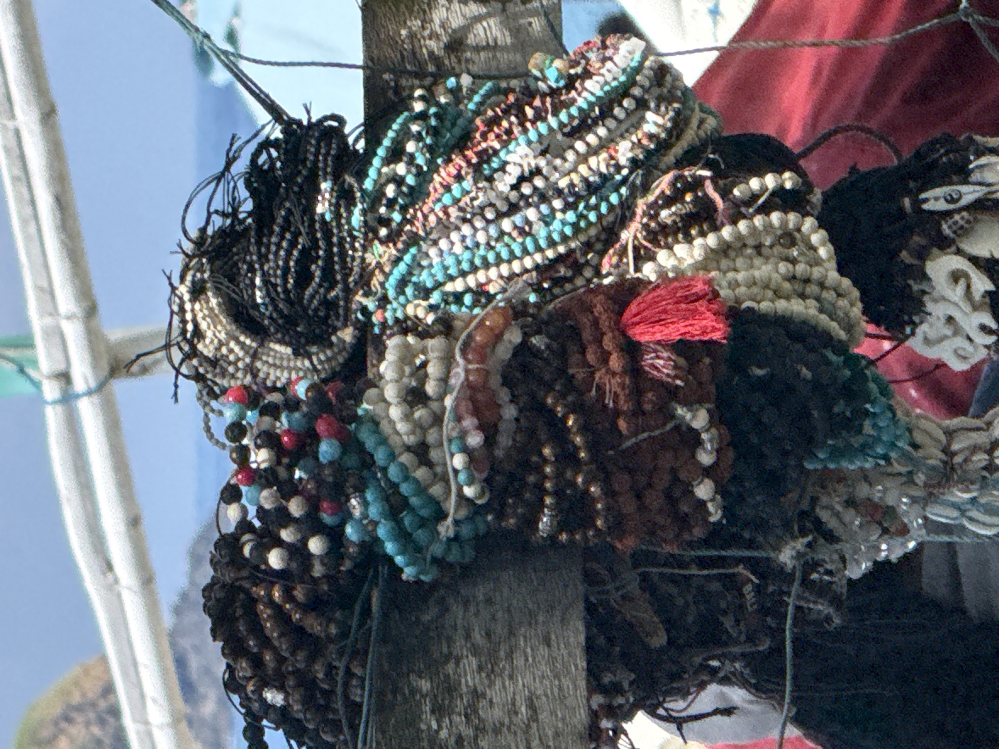 A collection of beaded necklaces hangs from a weathered wooden post. The beads are diverse in color, including black, brown, white, turquoise, and red. One necklace has a red tassel. The background shows a blurry blue sky and a distant landscape. A white, metal structure is visible to the left. The scene likely takes place in Karangasem, Indonesia, based on the image's prompt.
