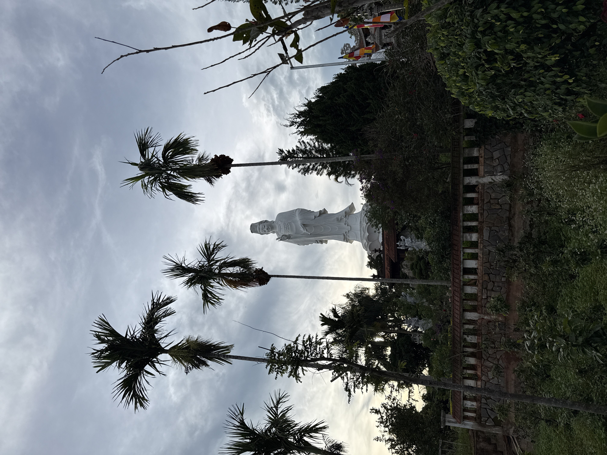 The image, taken in Thôn 5, Vietnam, features a white statue of a religious figure standing amidst lush greenery and palm trees under a cloudy sky. The statue is prominently positioned, overlooking a stone and brick wall. The environment includes various trees and plants, giving a sense of a garden or park setting. The sky is overcast, suggesting the time of day or weather conditions.