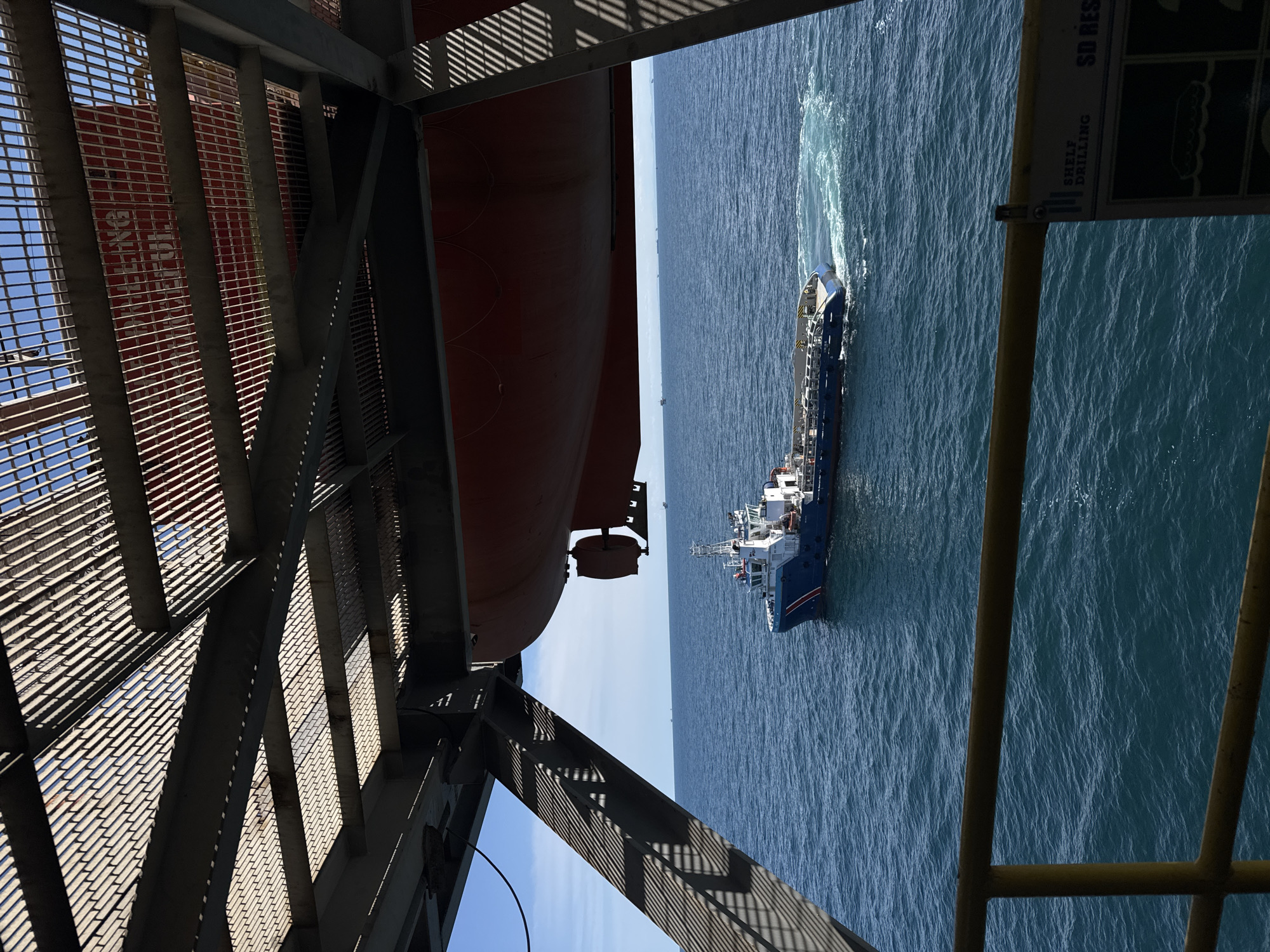A high-angle shot shows a blue and white vessel moving through blue ocean water, leaving a white wake behind it. The vessel is positioned to the right of the frame. To the left of the frame, a large red object, likely the hull of a ship or platform, is visible. This object is seen through a metal grate structure, casting striped shadows. Yellow railings are also visible in the foreground. A sign with text is attached to a yellow post. The text on the sign is partially visible and appears to read "SD RES" and "SHELL DRILLING". The sky is clear and blue. The lighting suggests it is daytime.