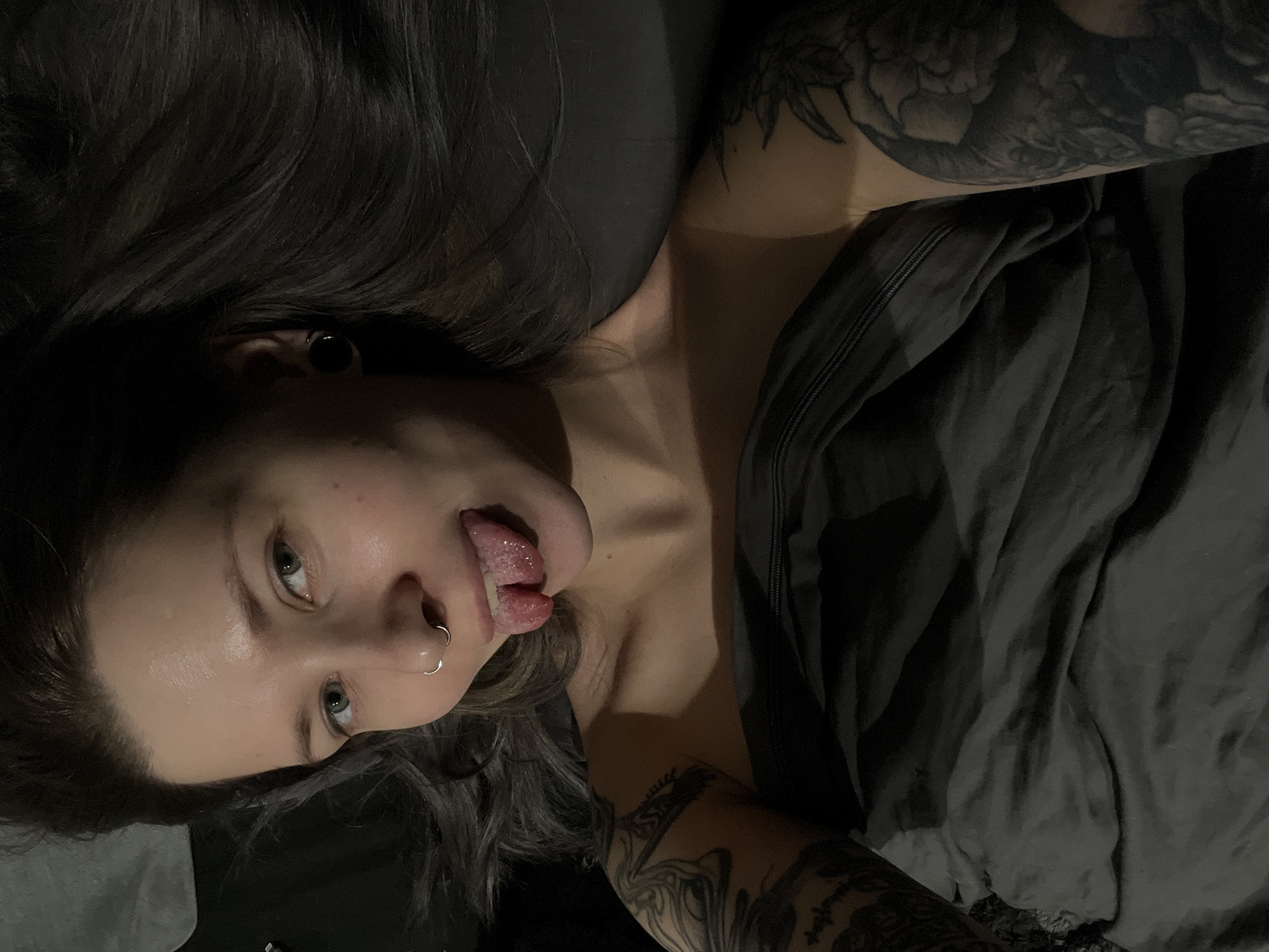 An indoor, eye-level shot captures a young adult person, likely female, lying down with their head angled towards the top of the frame. Their dark, long hair is spread around their head, blending into the dark background. The person is looking upwards with bright, light-colored eyes (possibly blue or green) and sticking their tongue out playfully.
Notable features include a silver hoop piercing on their left nostril and a black circular tunnel or plug in their left ear. Extensive black ink tattoos, depicting intricate floral or organic patterns, are visible on both arms and shoulders. The person's skin tone appears fair, with some redness on the cheeks and forehead, and a slight sheen suggesting moisture or oil.
They are partially covered by dark grey fabric, likely a blanket or bed sheet, which is rumpled and folded. A darker, possibly black, piece of clothing with a zipper is visible near their right shoulder and arm. The overall lighting is dim, suggesting it might be evening or night, creating soft shadows and highlighting facial features. The scene appears intimate and casual, possibly taken in a bedroom. There is no visible text in the image. The location context provided is Khimki, Russia, though no specific visual cues confirm this within the image itself.