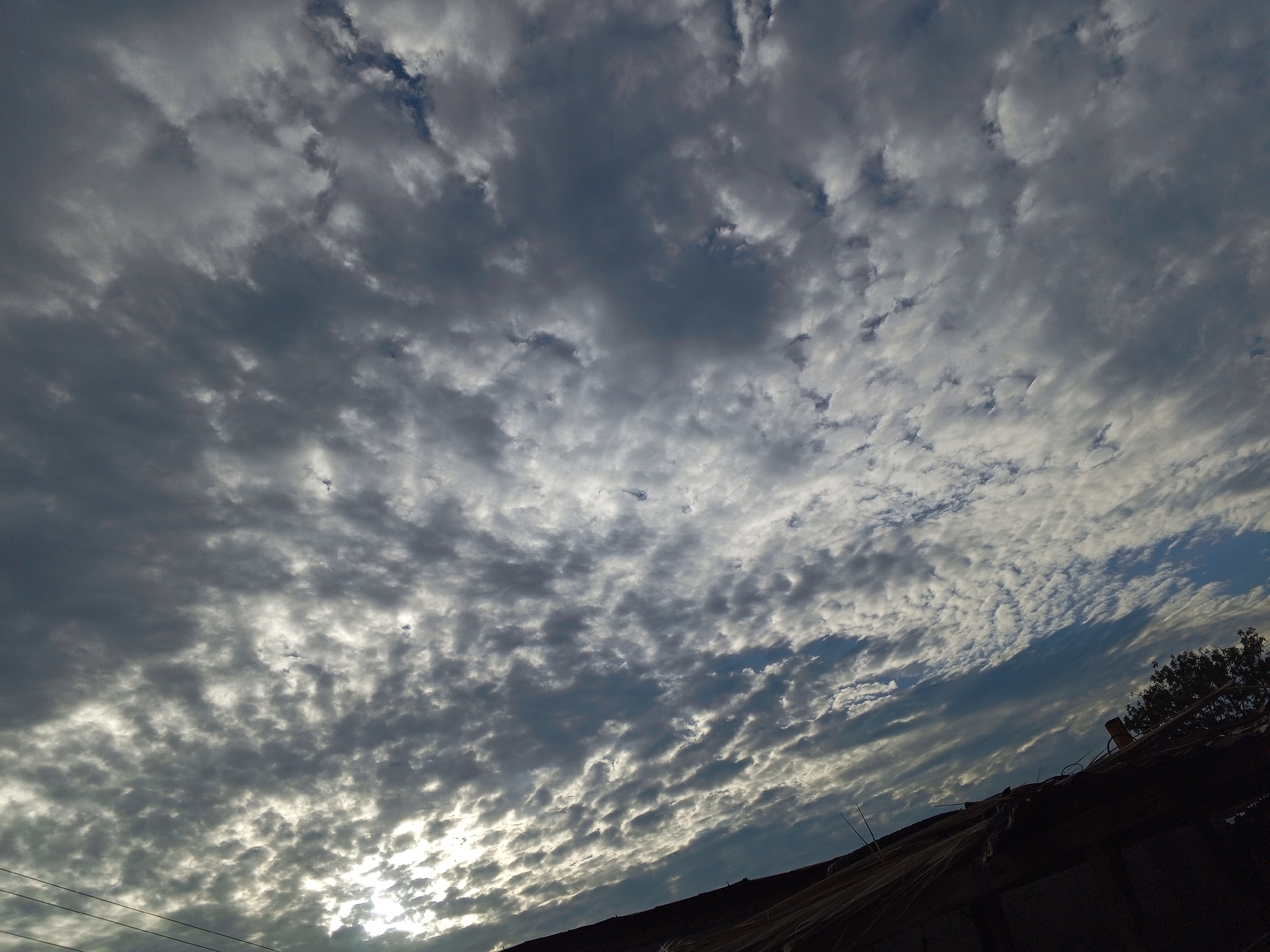 A low-angle shot depicts a cloudy sky over an unidentified structure. The sky is filled with various cloud formations, predominantly a mix of grey and white, with hints of blue sky peeking through in some areas. The clouds are dense and create a textured pattern across the frame. In the lower portion of the image, a dark, silhouetted structure with indistinct details can be seen. Its construction material appears rustic, possibly straw or thatch. A few tree branches are visible at the edge of the roof. The location is Unknown, Unknown.