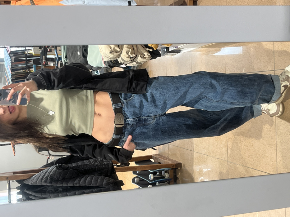 A person stands in front of a mirror, taking a selfie. They wear a light green crop top, baggy blue jeans with a black belt, a dark jacket, and tan-colored sneakers. The jeans' cuffs are rolled up. Visible is a silver necklace with a small pendant. In the background, clothing hangs, and skateboards are stored on a wooden rack below. The floor has beige tiles. The mirror's reflection shows a partial view of shelves and additional clothing.
