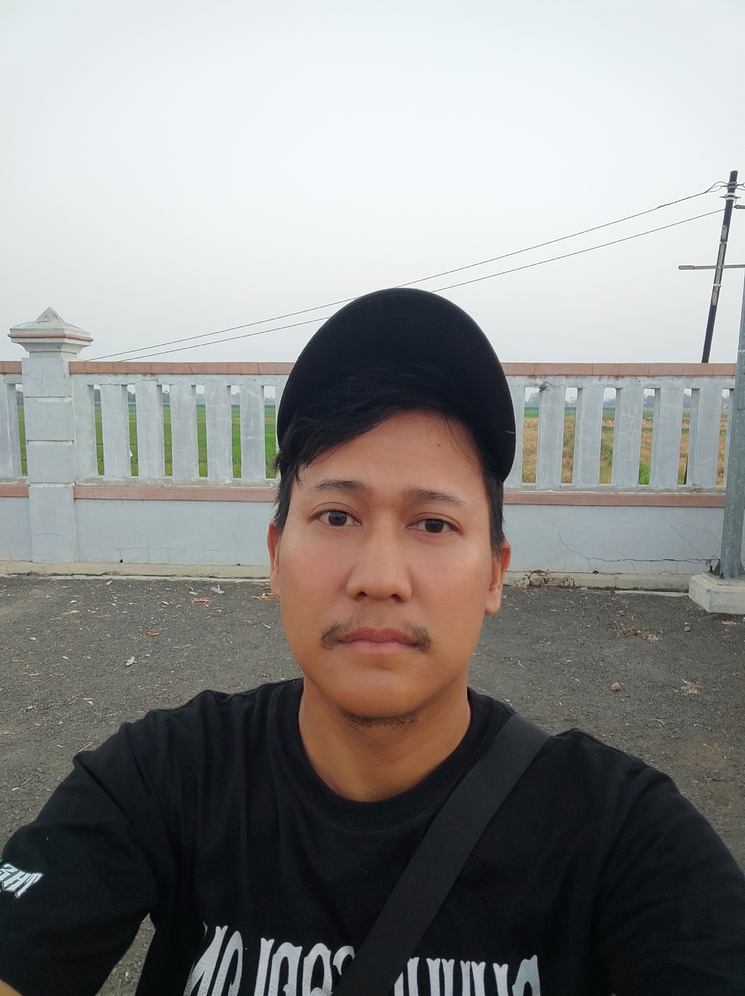 A man is taking a selfie outdoors in Pakusamben, Indonesia. He is wearing a black t-shirt and a black baseball cap. The man has a mustache and a short beard. In the background, there is a concrete fence with a pattern of vertical slats, behind which a green field can be seen. A utility pole with wires is visible to the right of the fence. The ground in front of the fence is covered in gravel. The sky is overcast.