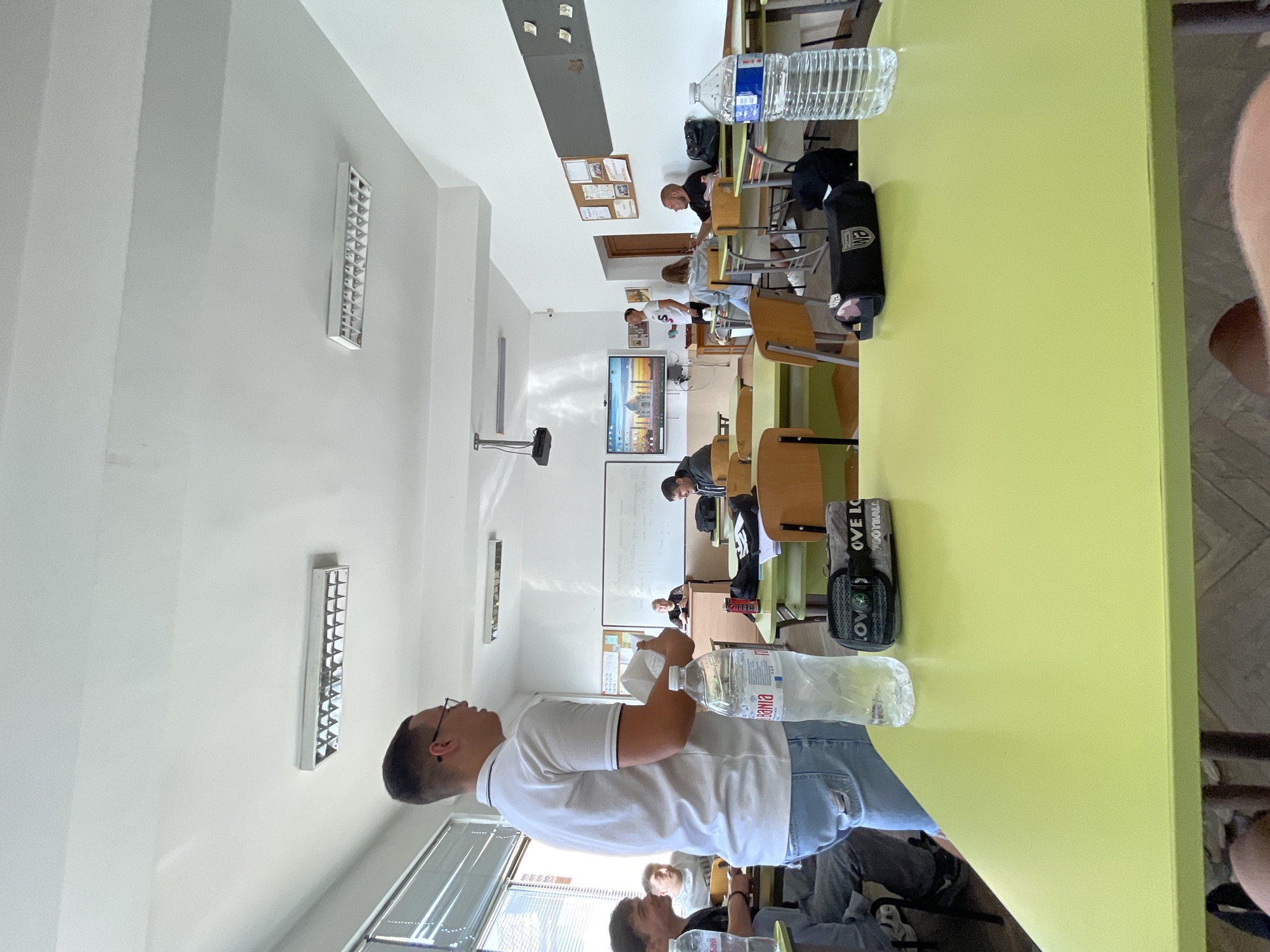 This image captures an indoor scene, likely a classroom or lecture hall, in what appears to be Zlatograd, Bulgaria. The room features light-colored walls, a white ceiling with multiple rectangular fluorescent light fixtures, and windows with blinds on the left side, indicating daytime.
In the foreground, a man wearing a white polo shirt, glasses, and blue jeans stands beside a large lime-green table, holding a plastic water bottle. His attention seems directed towards the middle of the room. To his right, another person is partially visible, seated at a desk.
Further back, several other individuals are seated at wooden desks and chairs arranged in rows. They appear to be students or attendees, with some looking towards the front of the room, suggesting a lesson or presentation is underway.
Key objects include numerous plastic water bottles on desks, one of which is clearly labeled "BILLA." A black pouch with the text "LOVE FOOTBALL" and a small compass attached to it is visible on a desk in the mid-ground. A projector is mounted on the ceiling, aimed towards the front of the room. At the front, there is a large digital display or television screen showing an image, possibly of the Taj Mahal, alongside a whiteboard. Information boards with various papers are pinned to the walls. The scene conveys a calm and focused atmosphere typical of an educational environment.