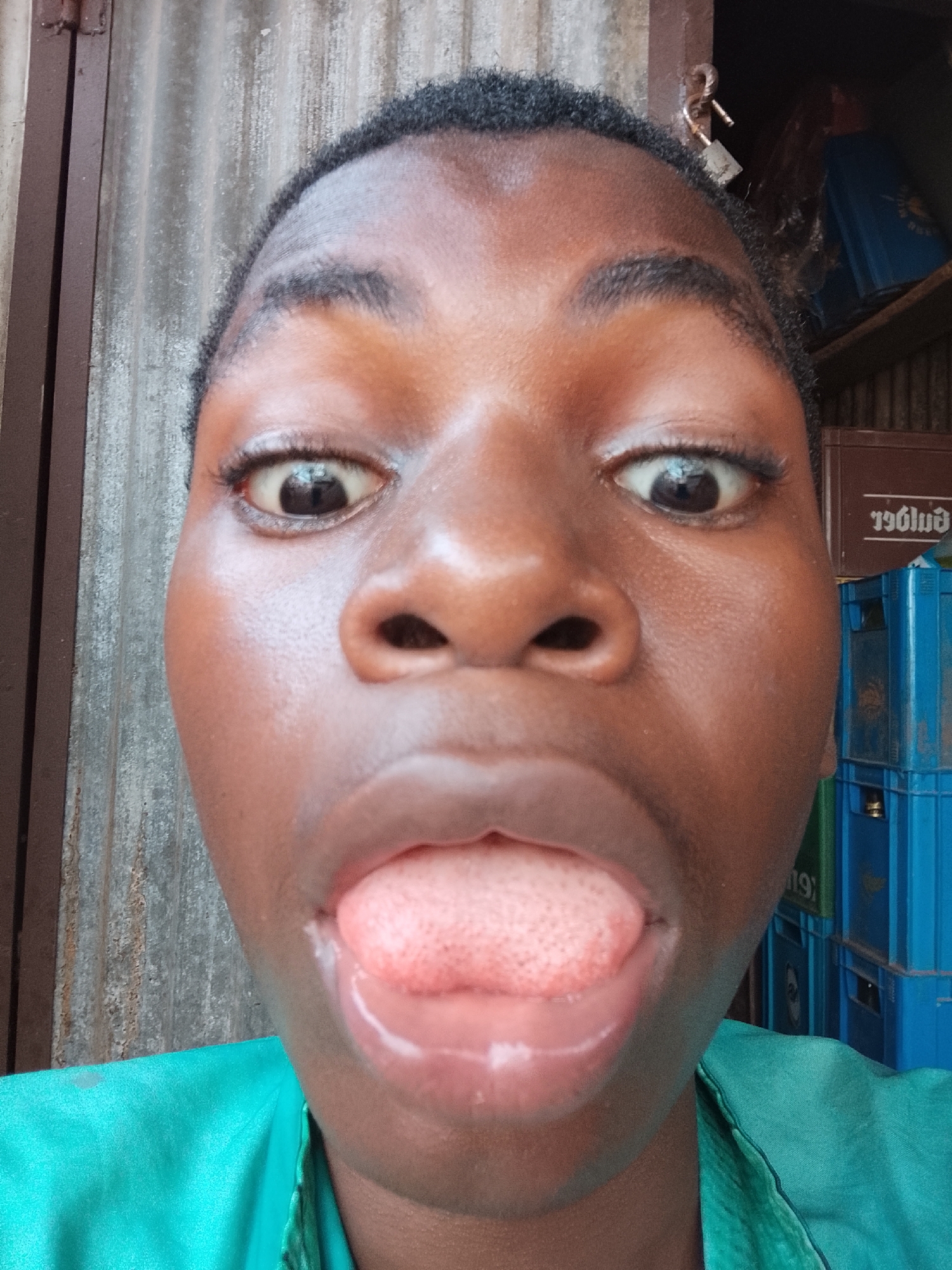 A close-up selfie of a young, dark-skinned person with short, dark hair and a bright green shirt is showing their tongue. The background is a corrugated metal wall to the left and stacked blue crates with beer advertising on the right. There is also a shelf behind the crates. The location is Nkpor, Nigeria.