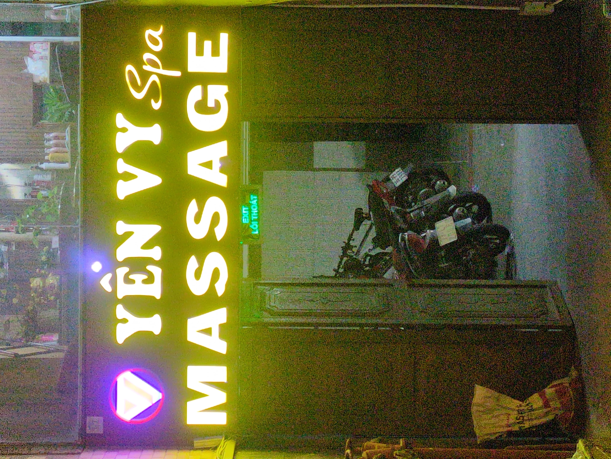 The image depicts a street scene in South Quy Nhon Ward, Vietnam. A neon sign for "Yến Vy Spa Massage" is prominently displayed. Behind the signage, an open doorway leads to an interior space where several parked motorbikes are visible. An exit sign with Vietnamese text ("EXIT LỐI THOÁT") is also present. An area containing various objects and materials is seen to the front and right of the doorway.