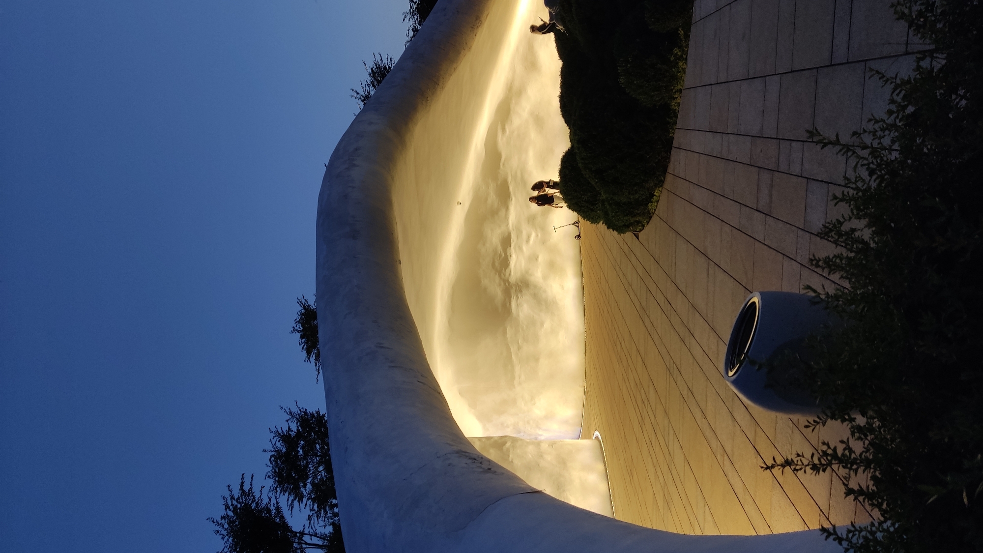 An outdoor scene at dusk shows a large, white, curved structure with an illuminated interior. The ground is paved with rectangular tiles. Several people are present, with two standing together in the illuminated area, one near a scooter. Another person is seated on the right. Lush green bushes line the curved edge of the paved area. A dark, modern-style trash receptacle is visible in the foreground. The sky above is a deep blue, indicating the late hour. The architectural style suggests a public space or park setting.