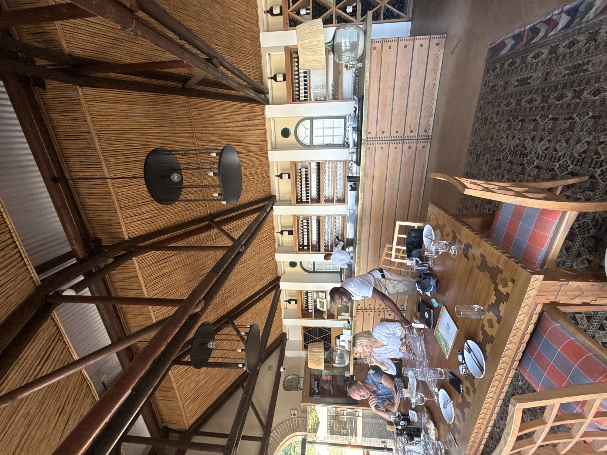 The image shows the interior of a restaurant located in Cape Town, South Africa. The restaurant features a high, peaked ceiling constructed of bamboo and dark wood beams. Several modern, black chandeliers hang from the ceiling. Along one wall are shelves stocked with wine bottles. A long, wooden bar runs adjacent to the wine shelves. At a large, ornately carved wooden table, a group of people are seated. A waiter, wearing a white shirt and khaki pants, is interacting with the diners at the table. The table is set with wine glasses, plates, and cutlery. The chairs have patterned cushions. A rug is visible in the background, adjacent to the wooden table. Through an arched doorway, some greenery is visible, indicating an outdoor setting beyond.