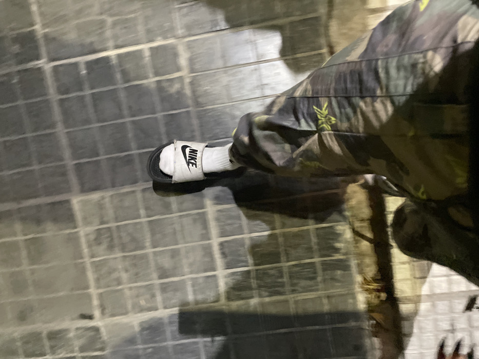 A first-person, downward-angle photograph captures a single individual's lower right leg and foot. The subject wears camouflage-patterned trousers, a white sock, and a black Nike slide sandal with white "NIKE" branding across the strap. The foot is positioned on a ground surface composed of gray, square tiles arranged in a grid pattern. Visible dirt and grime are present within the grout lines between the tiles. The tiled surface exhibits a slight reflectivity, suggesting moisture or a polished finish. Overhead illumination casts a defined shadow of the leg and foot extending to the left foreground. A partial, blurred view of a dark object with reddish-orange accents is visible in the lower-left corner of the frame. The immediate environment consists solely of the tiled ground, with no discernible landmarks or contextual elements to specify a location within the city of Buenos Aires, Argentina.
