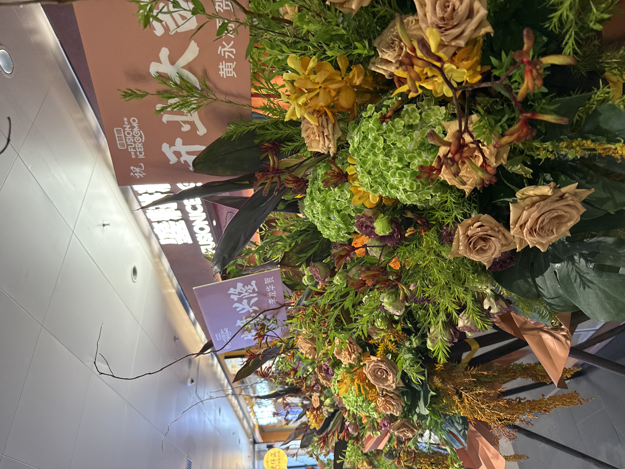 An indoor scene of a flower arrangement display with banners in the background. The flower arrangement features a variety of flowers and greenery in shades of brown, yellow, orange, green, and purple. The banners are in Chinese and feature text and logos. One banner is pink and has the words "FUSION ICEROOM" and Chinese characters that translate to "Congratulations on your opening." Another banner is purple and has Chinese characters that translate to "Flourishing and Prosperous" and a name. The scene appears to be in a commercial or public space, possibly a mall or event venue in Tianhe District, China, given the presence of the banners with Chinese text. The lighting suggests an indoor environment with overhead illumination. No people are visible in the image.