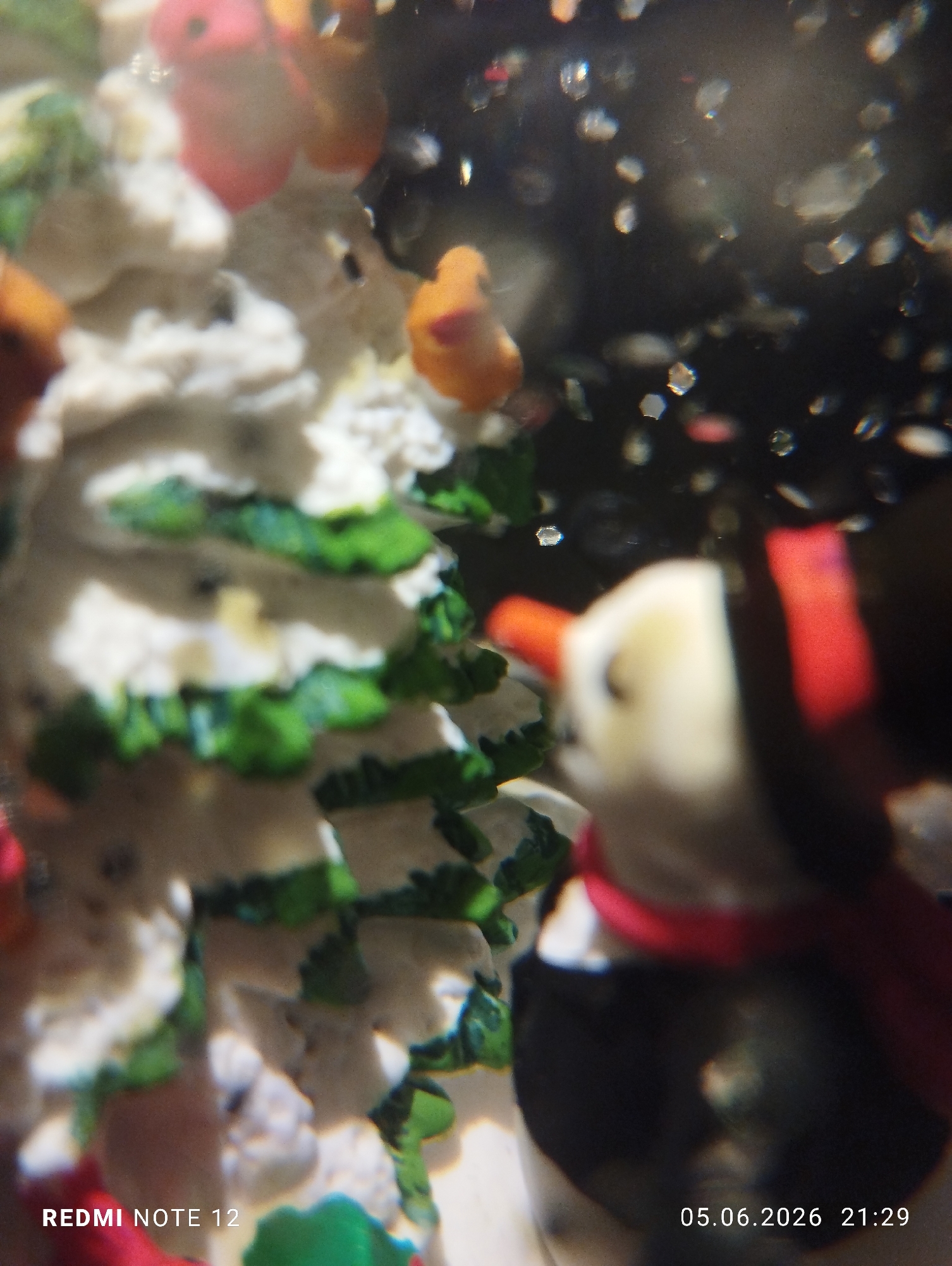 The vertical image presents a shallow depth of field close-up, likely of decorative figurines. In the immediate foreground, positioned to the right, a blurred figurine resembling a snowman is partially visible. It displays a white upper body with dark, circular eyes and a small orange detail indicative of a nose, a black lower body, and a red scarf. To the left, a portion of another out-of-focus decorative item, possibly a miniature Christmas tree, features white textured segments atop green elements. Throughout the midground and background, additional blurry forms in red and orange are discernible. The dark background is populated by numerous small, bright, shimmering specks, some exhibiting hexagonal forms, suggesting light reflections on particles in a liquid or dust. A watermark reading "REDMI NOTE 12" is located in the bottom left corner. The timestamp "05.06.2026 21:29" is present in the bottom right corner. The specific location in the city of Burgas, Bulgaria, cannot be determined from the image.