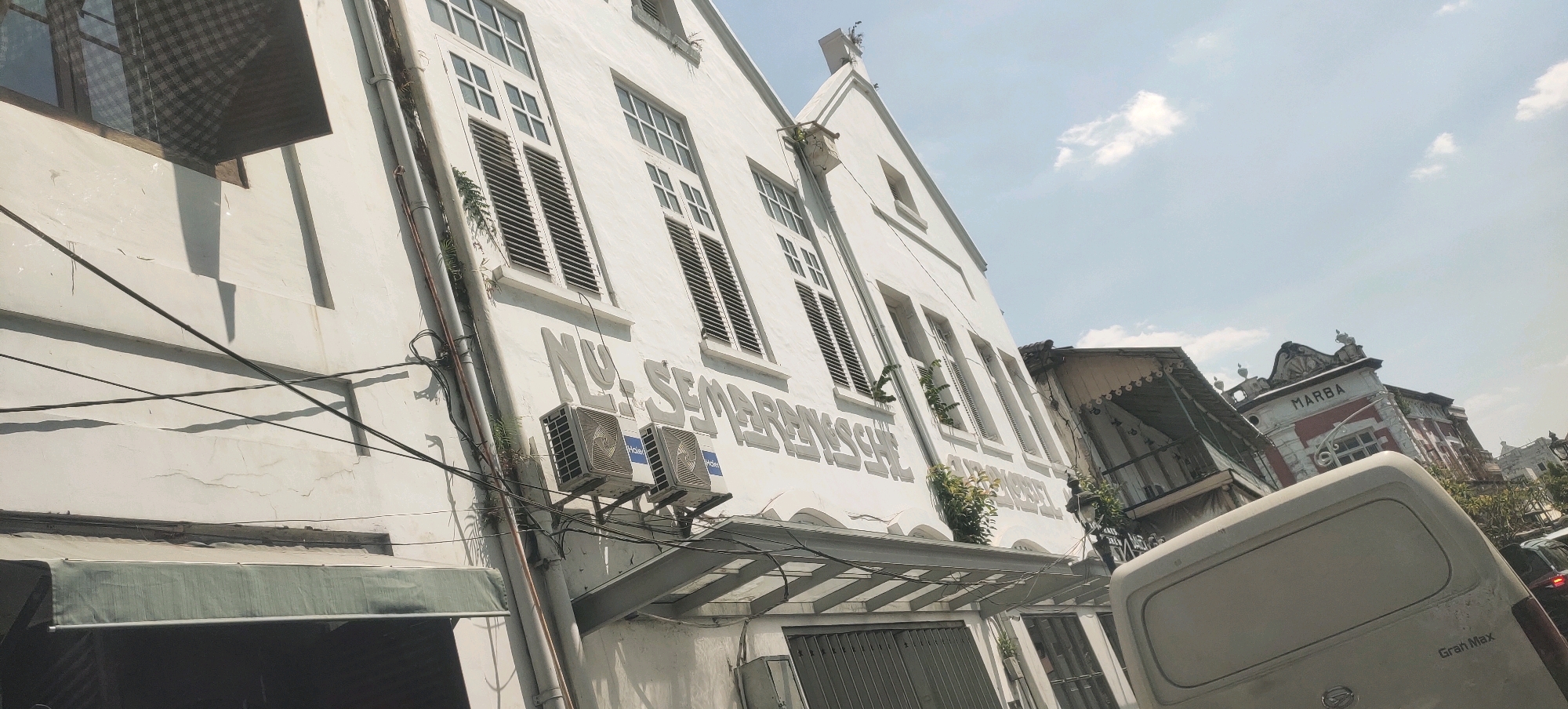 The image shows a street scene in Semarang, Indonesia, with a focus on a white, two-story building. The building has multiple windows with dark shutters and raised lettering that reads "NU. SEMARANG." Two air conditioning units are mounted on the exterior wall below the lettering. The building appears to be older, with some visible cracks and wear on the facade.
In the background, another building with red and white trim and the word "MARBA" is visible. A portion of a white van is in the foreground, obscuring the lower part of the scene. Power lines are strung across the front of the building. The sky is blue with scattered clouds. The overall impression is of a somewhat aged urban environment in a tropical climate.