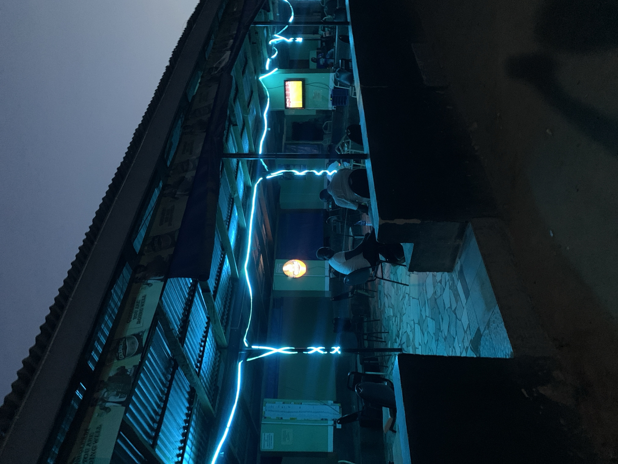 The image captures an evening scene at a casual establishment, likely an outdoor bar or cafe, situated near New Stadium in Nigeria. The setting is illuminated by prominent artificial light sources against a dark, possibly twilight or night sky.
The primary subjects include several individuals and the architectural elements of the venue. A building with a corrugated metal facade runs along the left side of the frame. Attached to this facade are banners, with visible text including "GOOD YOU ARE DOING WELL" and a partially visible "GOLDEN...". The building is topped with a dark roofline.
Inside the establishment, an open seating area is visible on the right. Several patrons are seated at tables. One person wearing a white shirt and dark cap is seen from behind, seated at a table in the foreground. Further back, another person in a checkered shirt is also seated. Their postures suggest a relaxed atmosphere, as they appear to be socializing or observing their surroundings.
The most striking visual element is the network of bright blue neon lights that traverse the space, creating a dynamic, squiggly pattern overhead and forming an "X.X" shape near the center. Additional light sources include an orange circular light fixture in the mid-ground and a rectangular screen or television emitting a yellowish glow further back. The ground is paved with patterned tiles in shades of light blue and grey. The overall mood is calm and inviting, typical of a relaxed evening gathering spot.