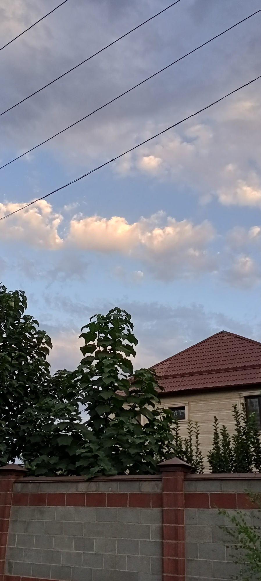 The image depicts a residential scene in Sokuluk, Kyrgyzstan. In the foreground, a brick and concrete fence lines the bottom of the frame. Behind the fence, lush green trees with large leaves are prominent, with a portion of a house visible through the foliage. The house has a light-colored facade and a reddish-brown tiled roof. The sky above is a mix of blue with scattered, soft-edged clouds, some of which are illuminated with a warm glow, suggesting it might be late afternoon or early evening. Several black power lines are stretched horizontally across the upper portion of the sky. There are no people or animals visible in the image. The overall impression is one of a tranquil, suburban setting.