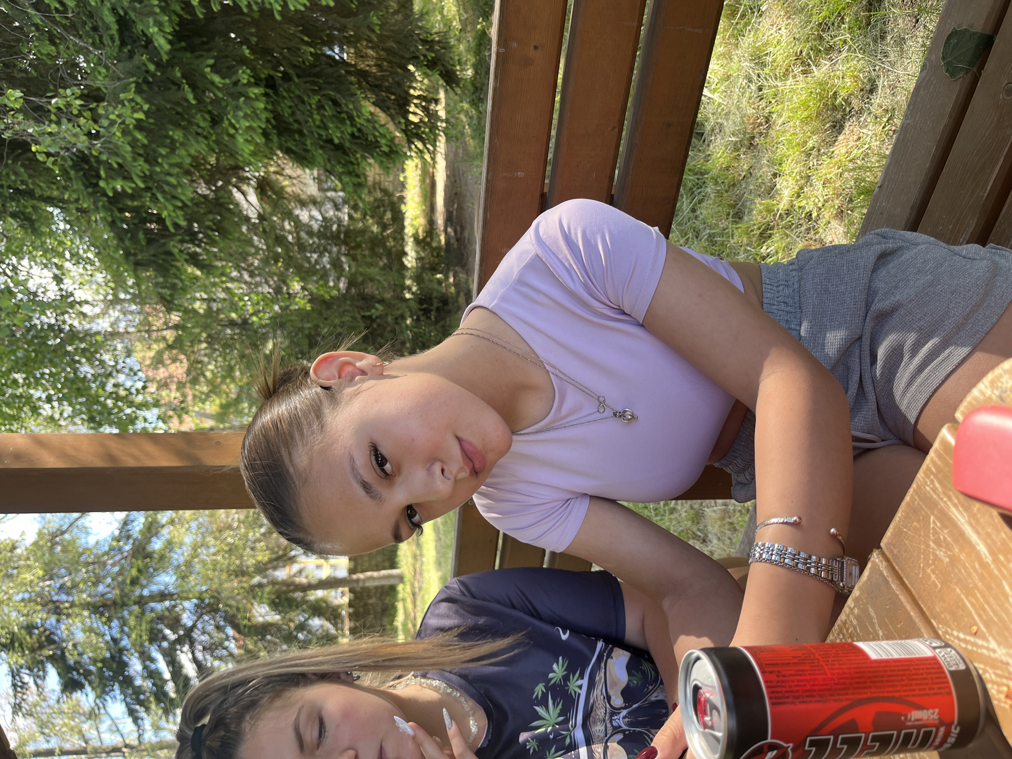 Two females are visible in an outdoor setting with vegetation in the background. One female wears a light purple top and grey shorts. She has her hair pulled back and is wearing a necklace, a watch and bracelets. The other female is wearing a dark colored t-shirt with a graphic print and a beaded necklace. A red can is on a wooden surface near them. The location appears to be Granitsa, Bulgaria, based on the image context.