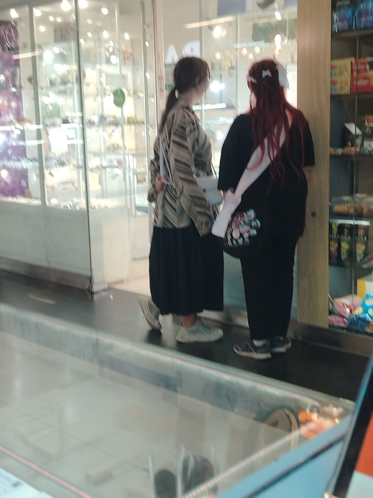 In a commercial space in Izhevsk, Russia, two individuals are observed from behind. One is wearing a striped jacket, a long black skirt, and light-colored sneakers. The second individual has red hair adorned with white ribbons, a black top, black pants, and dark sneakers; they also have a white sash across their body with anime characters printed on a tote bag. Both are standing in front of a store window displaying merchandise. To the right is a shelving unit stocked with packaged goods. The foreground shows a glass barrier. The overall setting suggests a mall or shopping center environment.