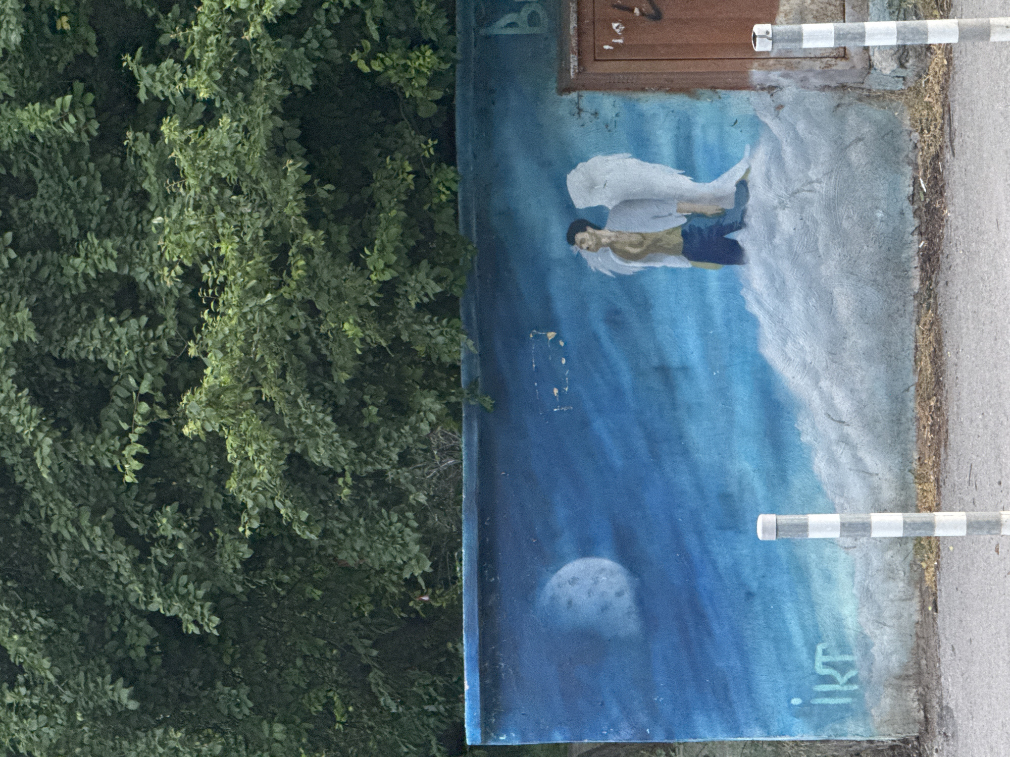 The image shows a mural on a concrete wall in Sofia, Bulgaria. The mural depicts an angel with white wings, standing on a cloud. The background is a blue sky with a moon. To the left of the mural, the word "IKT" is spray-painted. In the lower right corner, there is a brown wooden door. Two bollards with black and white stripes are visible in the foreground, one in front of the mural and another to the right of it. Overhanging the wall is dense green foliage from trees.