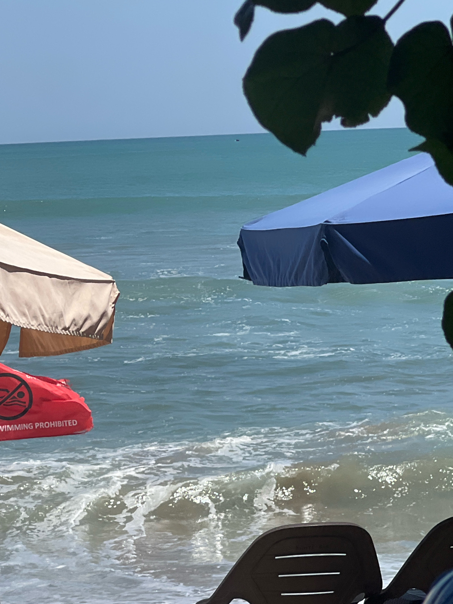The image depicts a beach scene with the ocean in the background. Two beach umbrellas are visible, one light beige and the other dark blue. In the foreground, a red float with a "no swimming" symbol and text reading "SWIMMING PROHIBITED" is partially visible. The ocean has moderate waves, and the sky is clear blue. The scene appears to be set in Kuta, Indonesia, as suggested by the context.