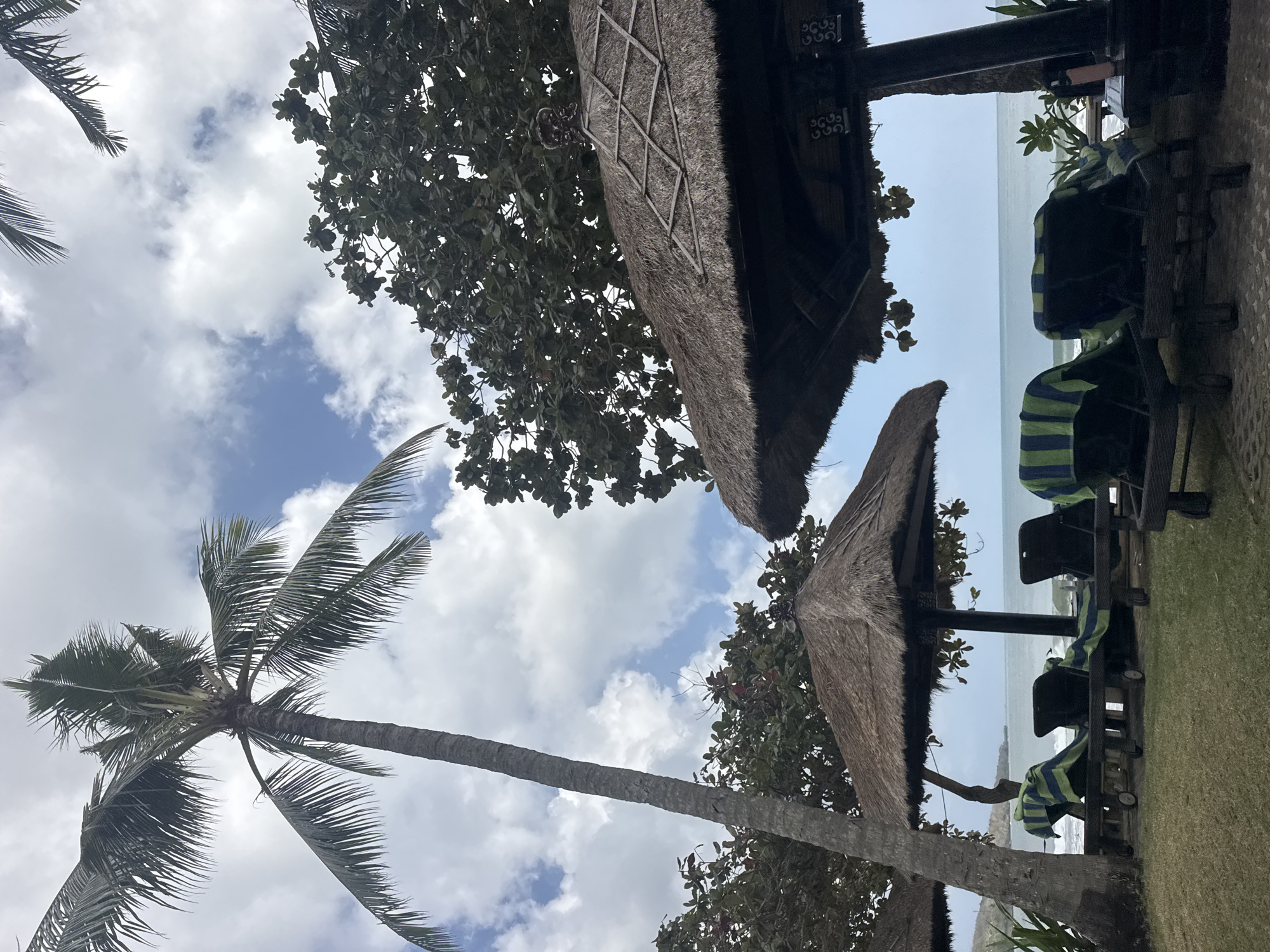 The image depicts a serene beach scene in Kuta Selatan, Indonesia, likely at a resort. In the foreground, several lounge chairs covered with blue and green striped towels are arranged on a grassy area. Behind the chairs, two thatched-roof umbrellas provide shade. A tall palm tree stands to the left, its fronds reaching towards a sky filled with fluffy white clouds against a backdrop of blue. In the mid-ground, lush green trees form a dense canopy. The background reveals a calm ocean with gentle waves, meeting a distant shoreline. The overall atmosphere is tranquil and inviting, typical of a tropical vacation destination.