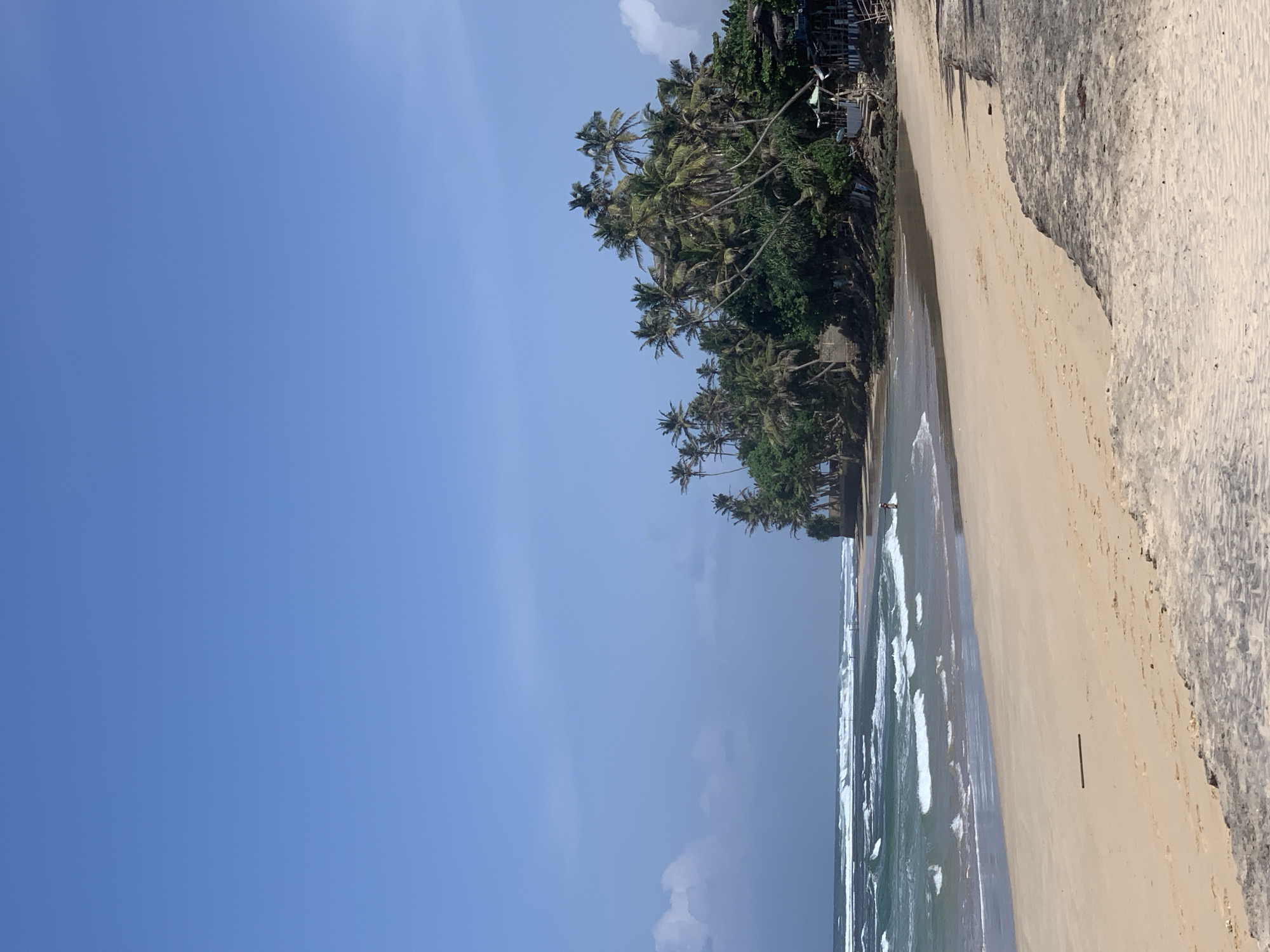 The image depicts a tropical beach scene in Kathaluwa, Sri Lanka. The foreground is dominated by a wide, sandy beach with a stone embankment on the right. The sand shows some rippled patterns from the wind. A thin, dark stick lies horizontally on the sand in the mid-ground.
Further back, the ocean meets the shore with white-crested waves rolling in. The water transitions from a deep teal to a lighter turquoise near the shore. A solitary figure, appearing to be a person, is visible in the water near the shoreline, possibly engaged in swimming or surfing.
The background features a lush line of palm trees and dense green foliage that extends along the coast. Several buildings, appearing to be small structures or huts with thatched roofs, are nestled amongst the vegetation on a slightly elevated area. The sky above is a clear, vibrant blue with a few wispy clouds on the left side of the frame. The overall impression is of a serene, coastal landscape under a bright, sunny sky.