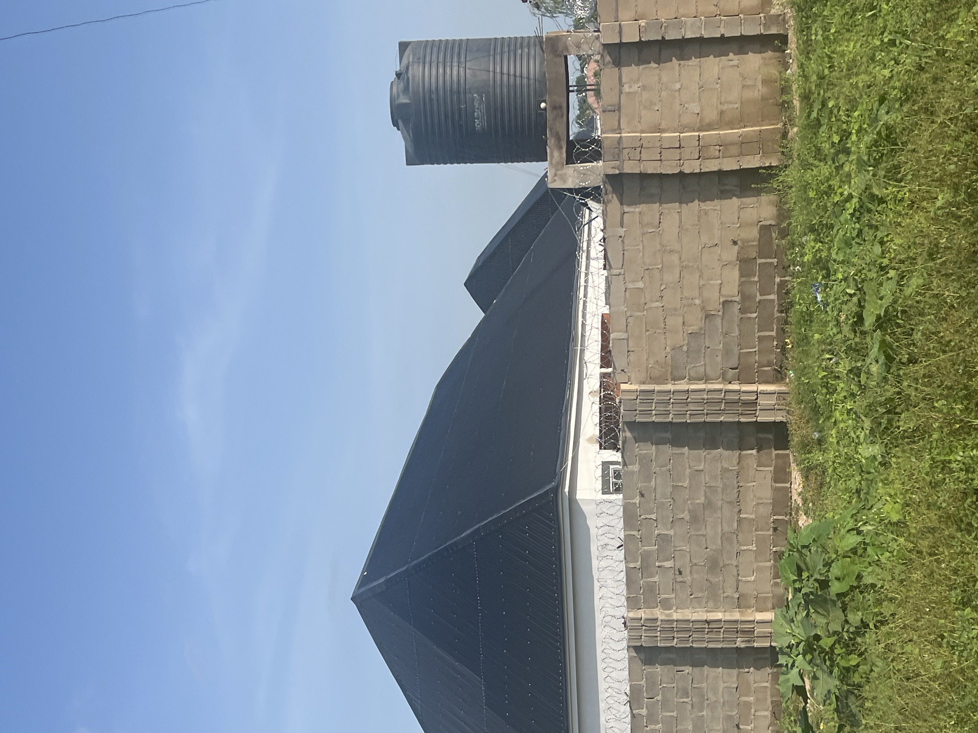 The image shows a residential property with a grey brick wall in the foreground, topped with barbed wire. Behind the wall, a white building with a dark, pyramid-shaped roof is visible, along with a large black water tank on a concrete platform. The sky is clear and blue. The environment is predominantly green with tall grass and foliage in front of the wall. The location is identified as Jalingo, Nigeria.
