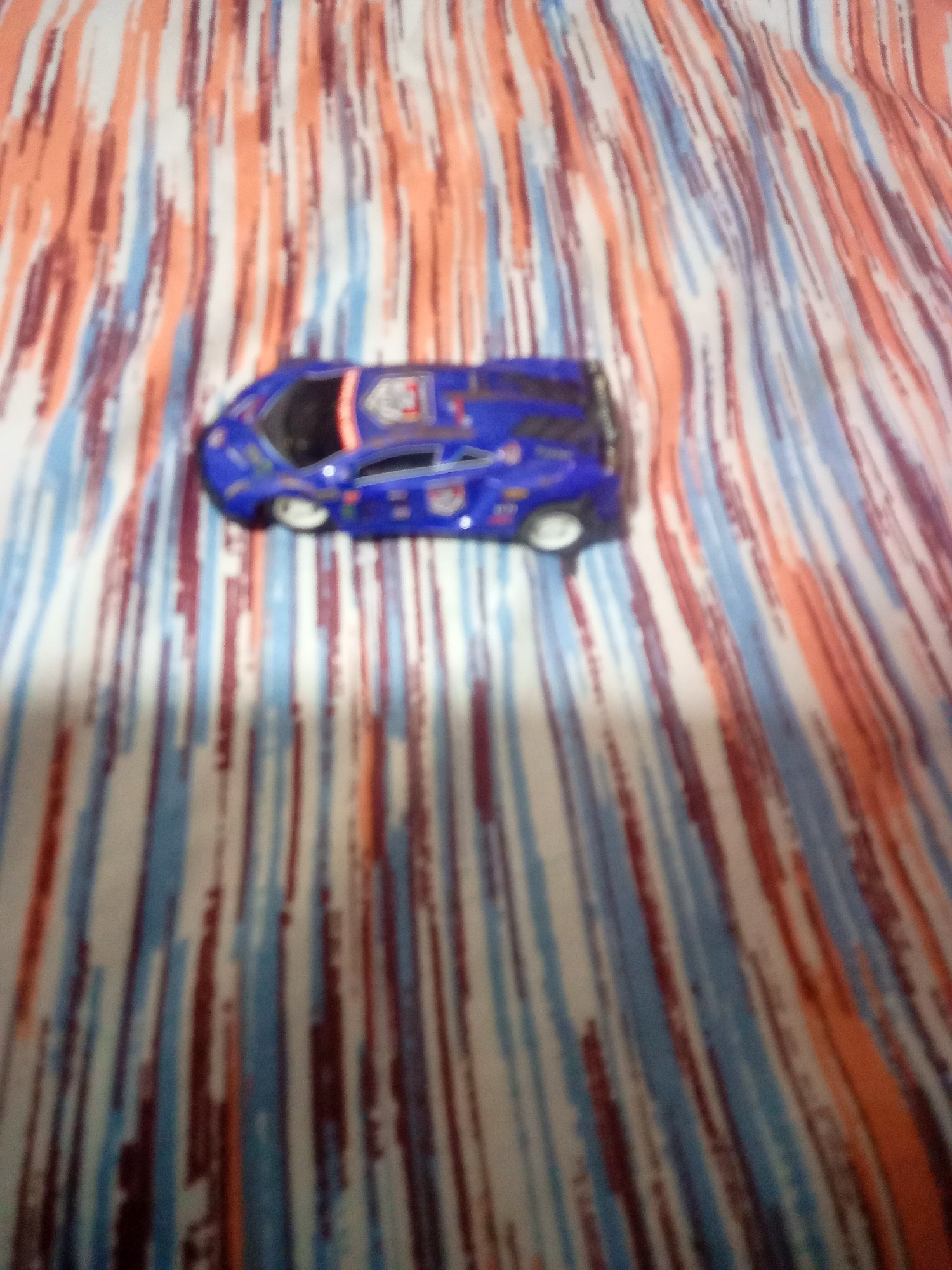 The image shows a blurry, blue toy car with white wheels placed on a patterned fabric. The fabric has vertical stripes in shades of orange, white, blue, and brown. The image is out of focus, making it difficult to discern fine details of the car or the fabric pattern. There is no indication of the city of La Tagua, Venezuela, or any other specific location. The scene depicts a static object on a textile surface.