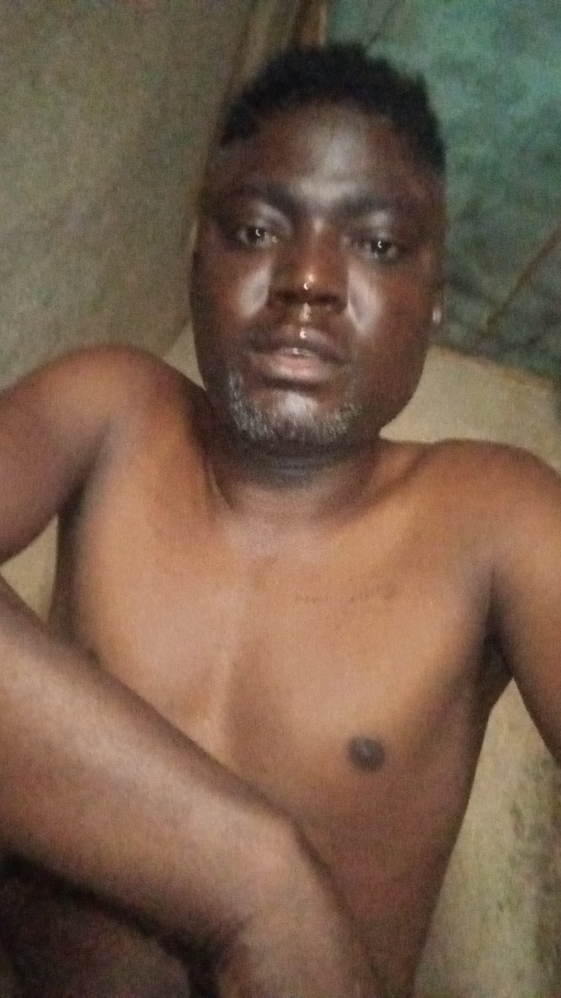 A man with dark skin and a salt-and-pepper beard is shirtless and looking directly at the camera. He has short, dark hair and is indoors. The location is Unknown, Unknown. The image has a grainy quality.