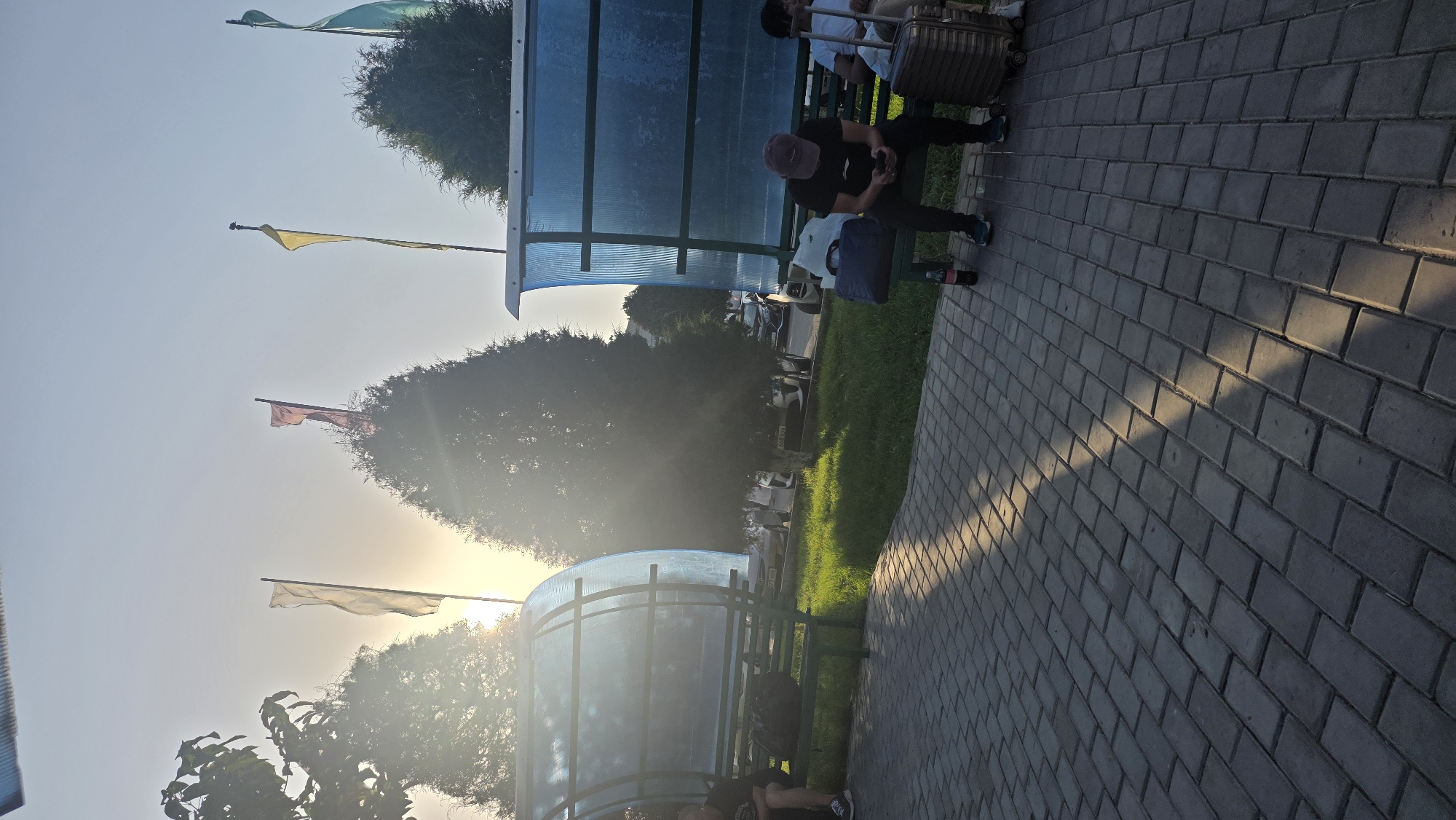 The image depicts a street scene at what appears to be a bus stop or waiting area in Nariman, Kyrgyzstan. The time of day is late afternoon or early evening, with the sun low in the sky, casting long shadows across the paved walkway. Three flags are visible in the background, one yellow, one red, and one greenish-blue, attached to poles atop trees.
In the foreground, a gray brick walkway is visible, with sunlight creating a diagonal pattern of light and shadow. A green metal bench is situated beneath a translucent blue and green shelter. On the bench to the left, a person is seated, facing away from the camera, with a dark bag placed beside them. To the right, another person, wearing a dark cap and black t-shirt, is seated on the bench, looking at their phone. A dark blue bag and a bottle of soda are placed on the bench next to them. A large, dark suitcase with wheels is positioned on the ground to the right of the second person. Another person is partially visible on the far right, sitting behind the suitcase. Behind the shelter, a grassy area is visible, with several trees. Further in the background, cars are parked along a street, and more trees and buildings can be seen. The sky is clear and bright, with the sun partially obscured by a tree.
