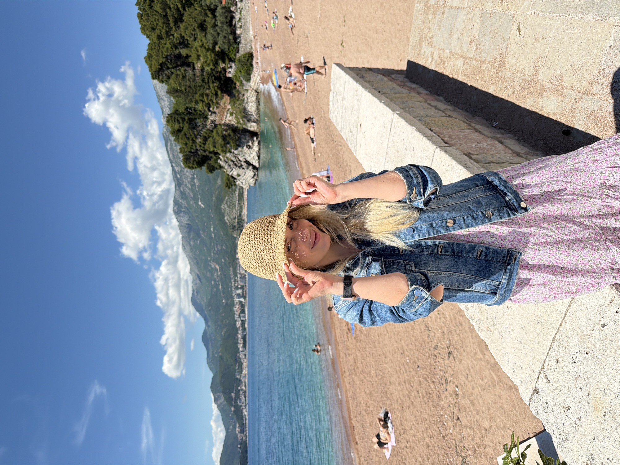 The image shows a woman in a straw hat and denim jacket posing for a picture. She is on a stone ledge or wall, overlooking a beach with people, and the Adriatic Sea in Sveti Stefan, Montenegro. The backdrop includes mountains and a blue sky with some clouds.