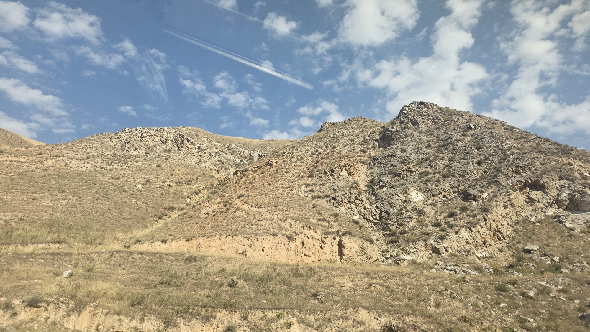 A low-angle, wide shot captures a dry, rocky mountain landscape under a blue sky dotted with white, wispy clouds. The foreground features dry, yellowed grass covering a gently sloping terrain, with evidence of erosion forming terraces and gullies. The midground is dominated by the imposing, rocky slopes of the mountain, characterized by a mix of brown and grey rock formations sparsely covered with dry vegetation. The upper portion of the image reveals the mountain's peak, a jagged crest against the sky. A faint, white contrail is visible in the upper left quadrant of the sky. The scene is devoid of human presence or any artificial structures, presenting a natural, arid environment. The lighting suggests it is daytime, with sunlight illuminating the terrain. This landscape is characteristic of the mountainous regions surrounding Taldyk, Kyrgyzstan, where such arid, rocky terrains are common.