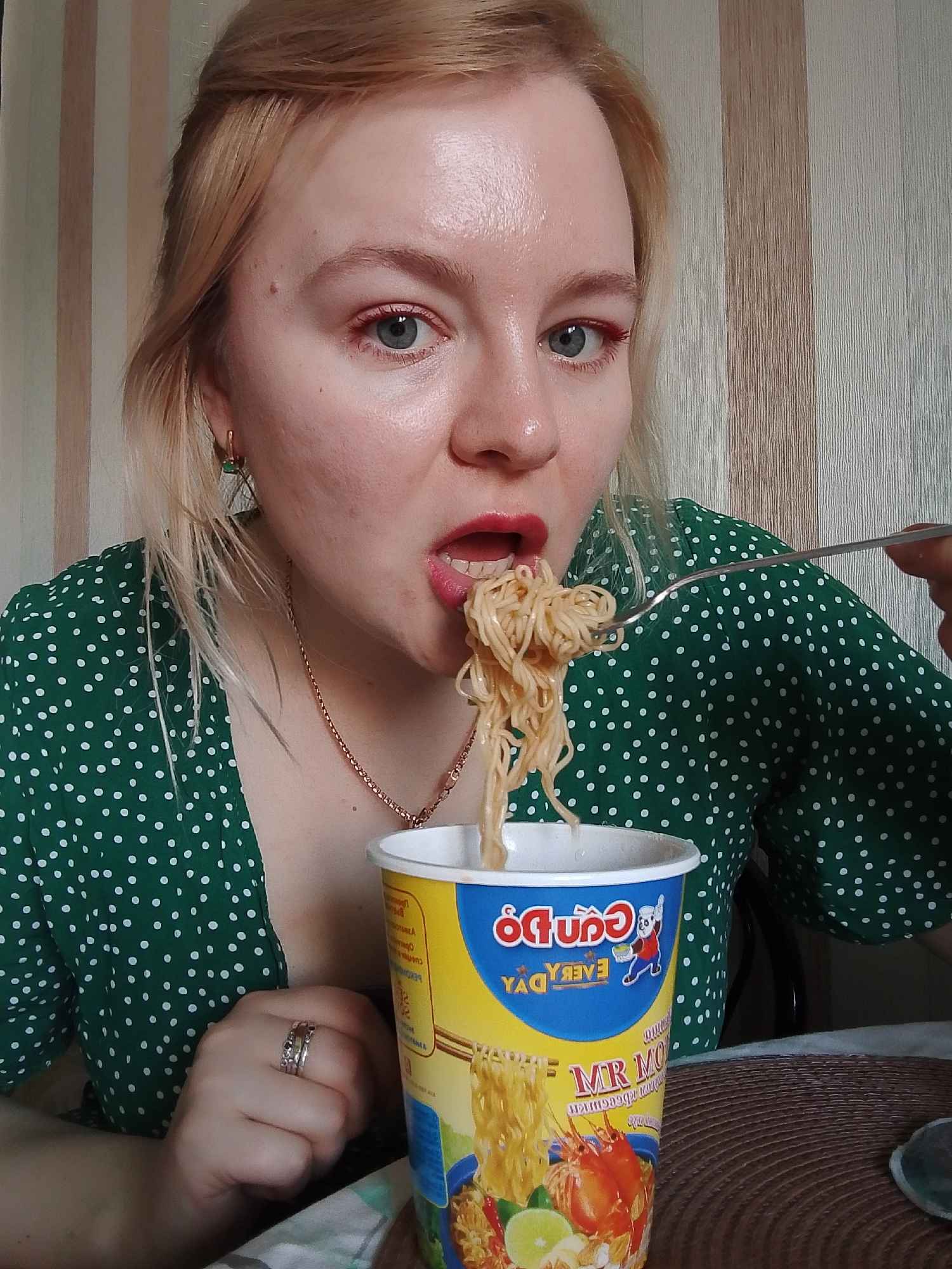 A young woman with blonde hair and fair skin is eating noodles with a fork indoors. She is wearing a green dress with white polka dots and has red eyeliner and lipstick. She is looking at the camera with her mouth open and noodles on the fork being brought to her mouth. She has a gold chain around her neck and rings on her left hand. A cup of Vietnamese instant noodles, brand name "Cung Do," is on the table in front of her. The table has a woven brown mat on it. The background is a wall with vertical beige stripes. It is not possible to determine the location in Izhevsk, Russia from this image alone.
