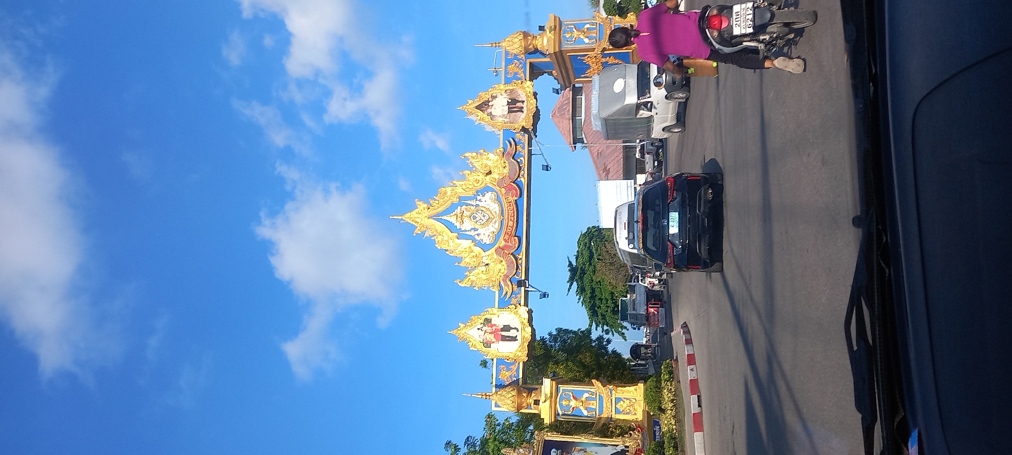 The image, taken from within a vehicle, captures a bright daylight street scene in Thong Sala, Thailand. A grand, ornate archway, primarily gilded in gold with blue accents and traditional Thai architectural elements, spans across the road overhead. This ceremonial archway features intricate carvings, pointed spires, and flame-like motifs. Framed portraits are integrated into the archway's structure; one clearly shows a man and a woman in formal attire, likely members of the Thai royal family, while another depicts a single male figure in uniform.
Below the archway, an asphalt road carries traffic. A person wearing a purple shirt is seen riding a motorcycle on the right side of the road. The motorcycle's license plate displays "2กก", "6212", and "Thailand". Further along the road, several vehicles are visible, including a black SUV with a license plate reading "ภฉ 8877", and a white van. The road's edge features a curb painted with red and white stripes.
The sky is a vibrant blue with scattered white clouds, suggesting clear weather conditions. Green trees and foliage are present alongside the road and behind parts of the archway. In the background, a building with a reddish-brown roof can be seen. The bright sunlight indicates the photo was taken during the day, likely morning or afternoon. The overall scene depicts typical daily activity on a thoroughfare, marked by the distinctive cultural architecture of Thailand.