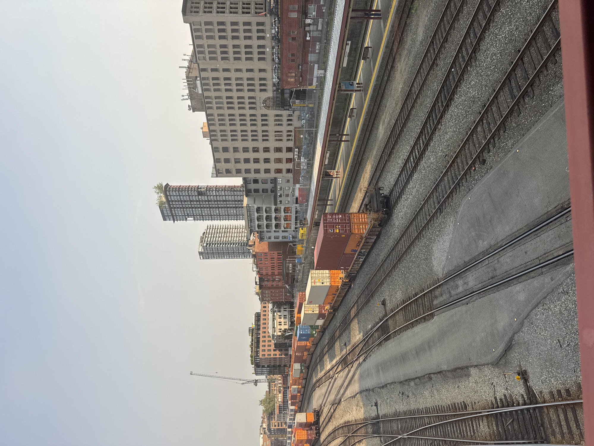 This is a high-angle, eye-level, medium shot of a busy railyard in the Gastown district of Vancouver, Canada. In the foreground, multiple sets of train tracks run parallel to each other. Several long freight trains are parked on the tracks, loaded with shipping containers of various colors, including orange, white, blue, and red. The trains stretch out into the middle ground, where they are juxtaposed with train platforms and low-rise brick buildings. In the background, a cluster of modern high-rise buildings dominates the skyline, with one particularly tall skyscraper featuring a green rooftop garden. A construction crane is visible in the distance, to the left of the tall buildings. The sky is clear and light blue, with the sun casting long shadows, indicating it is likely late afternoon. The overall scene depicts an active industrial and urban environment.