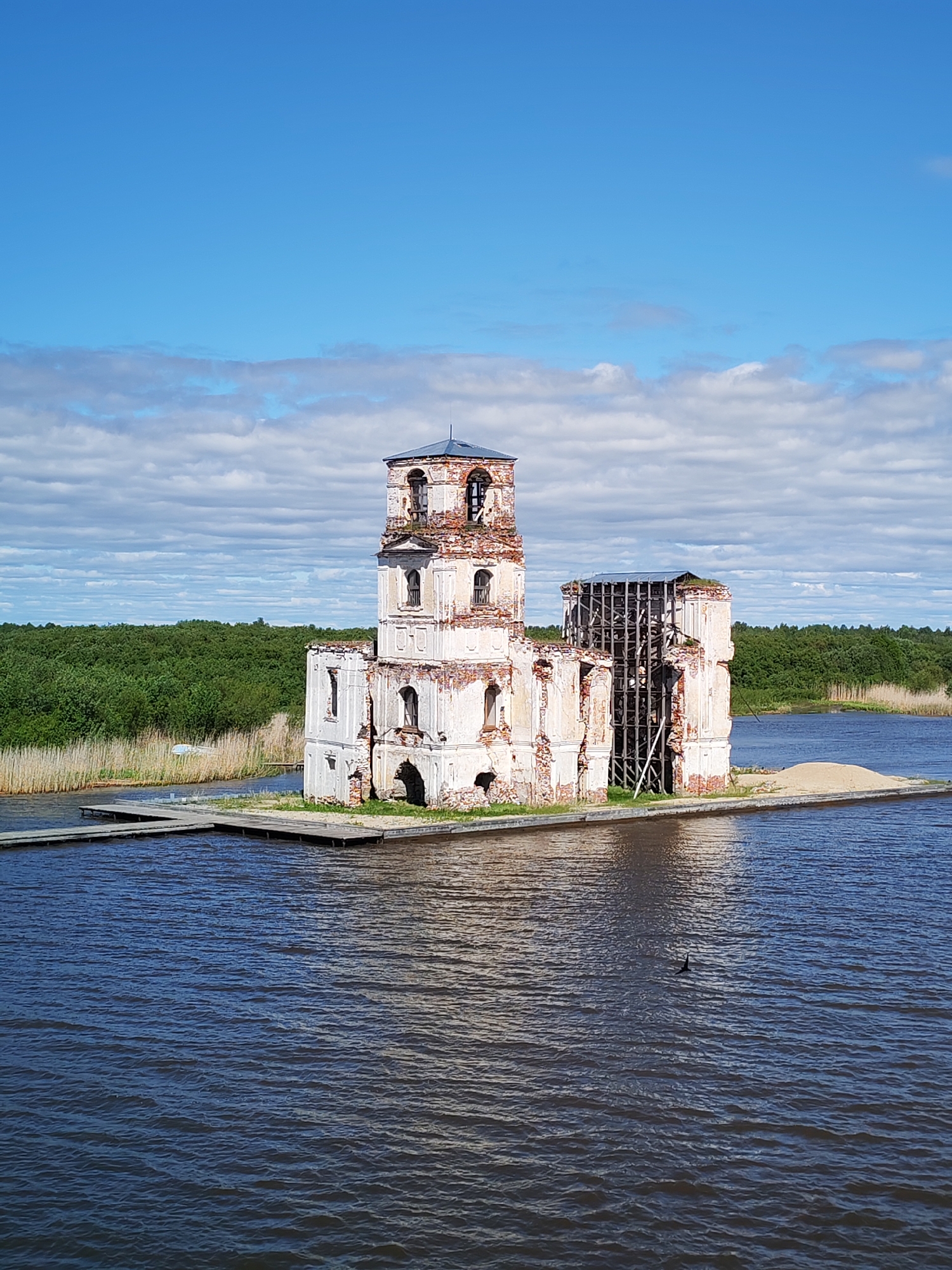 The image captures the partially ruined Church of the Nativity of Christ in Krokhino, Russia, standing on a small artificial island in the waters of a lake. The church is a multi-tiered structure of white brick with visible deterioration and areas missing. Scaffolding supports a portion of the building. A pile of sand sits adjacent to the church. A narrow wooden pier extends from the island towards the shoreline. A body of water surrounds the church. Beyond the water lies a strip of land covered in dense green foliage. The sky is a clear blue with scattered white clouds.