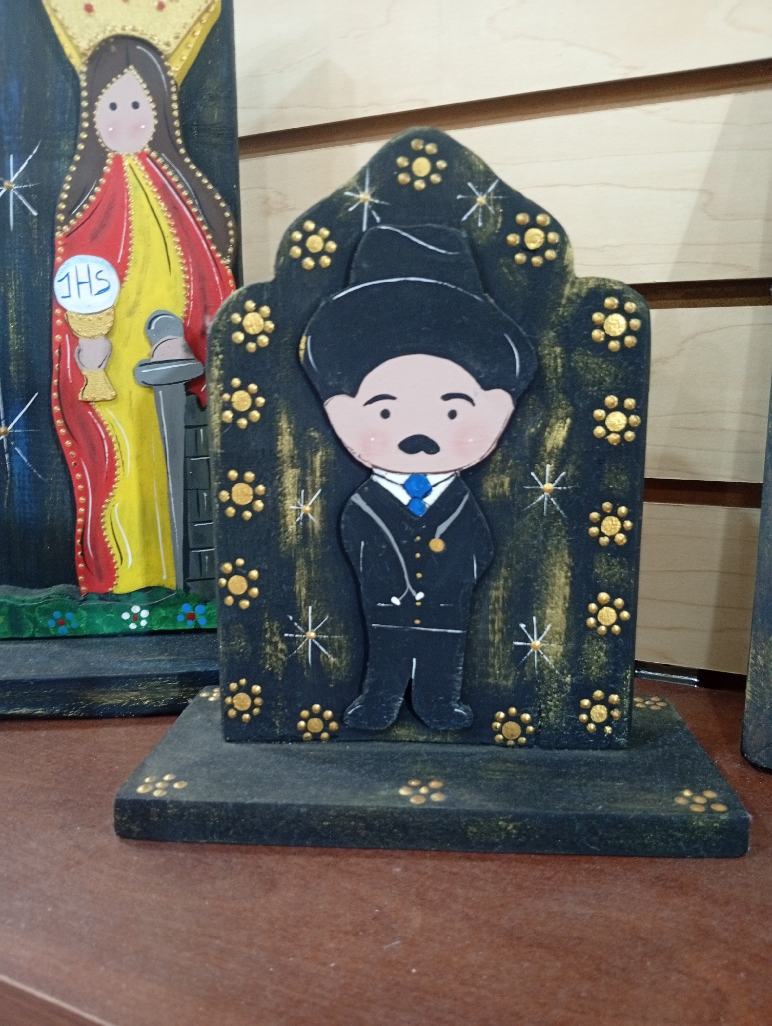 This is a close-up, eye-level, indoor shot of two folk art figures displayed on a wooden surface. The figure on the left is a tall, vertically oriented cutout of a woman, likely a religious figure, with a crown, red and yellow robes, and holding a white disc with "JHS" written on it. This figure is dark blue, with gold and red accents. To the right is a smaller, freestanding cutout of a man in a black suit with a blue tie and black hat. He is standing on a black base decorated with gold dots and starbursts. The background consists of horizontal wooden planks. The figures appear to be made of painted wood or similar material. No specific location within Juan Griego, Venezuela is identifiable from the visual information provided.
