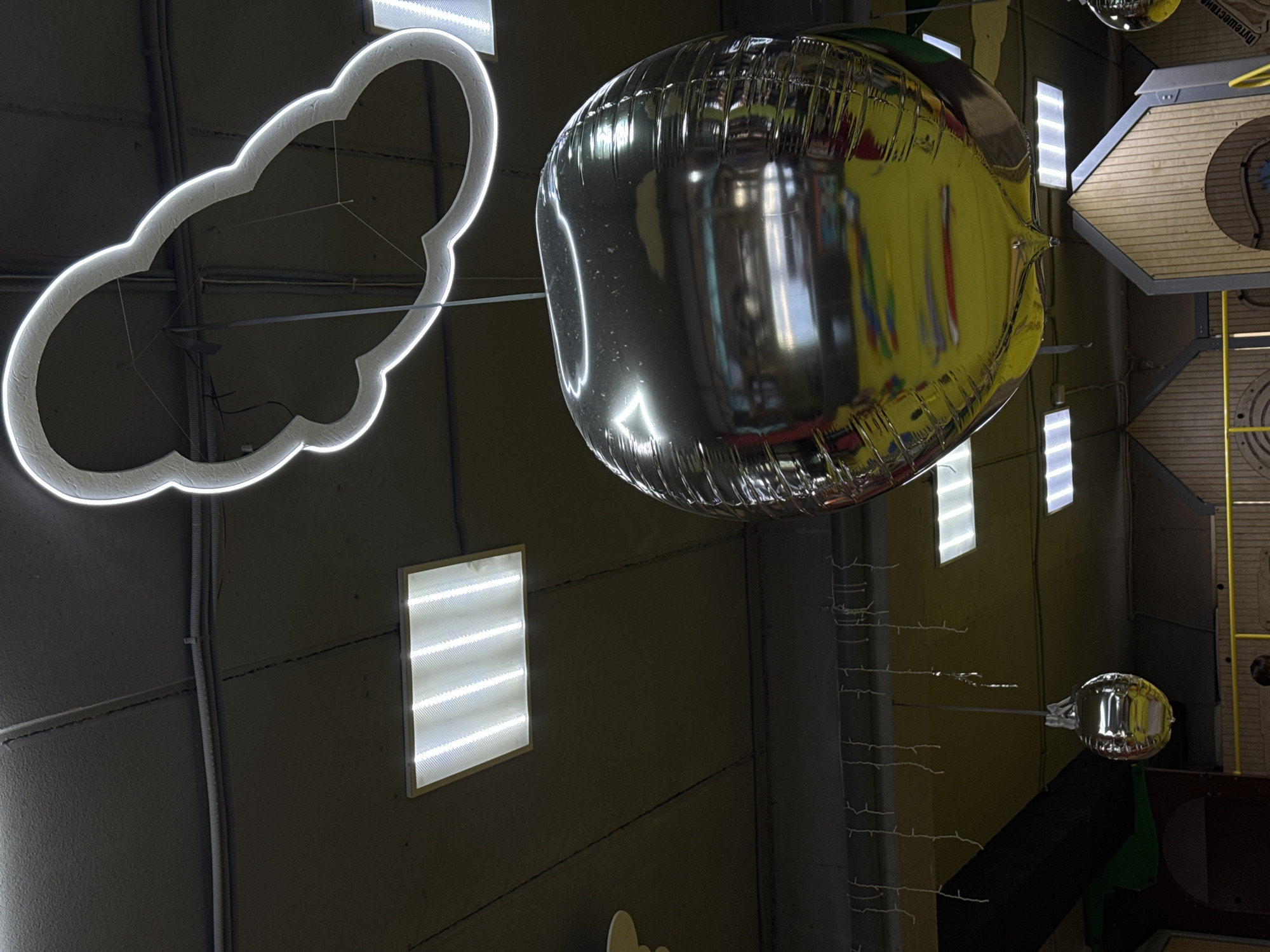 This is a medium-shot, indoor image taken in Mikhaylovsk, Russia, likely in a children's play area or entertainment venue, given the decor. The scene is dominated by the ceiling, which is a dark, textured surface with visible cracks and conduit piping.
Suspended from the ceiling are several illuminated elements. A prominent, cloud-shaped outline made of white light hovers in the upper portion of the frame. Below it, a large, spherical, metallic balloon, appearing to be silver or chrome, hangs by a string. The balloon is highly reflective, mirroring the yellow ceiling and colorful elements of the environment below. Its surface shows texture from its construction.
Other features visible on the ceiling include two rectangular fluorescent light fixtures, emitting bright white light, positioned on the left and right sides of the frame. Additionally, a series of small, white string lights hang downwards, resembling icicles.
In the background, partially visible structures with wooden paneling and arched openings suggest playhouses or similar activity areas. The overall lighting is a mix of artificial sources, creating a brightly lit yet somewhat dim environment due to the dark ceiling. No people are clearly visible in the frame.