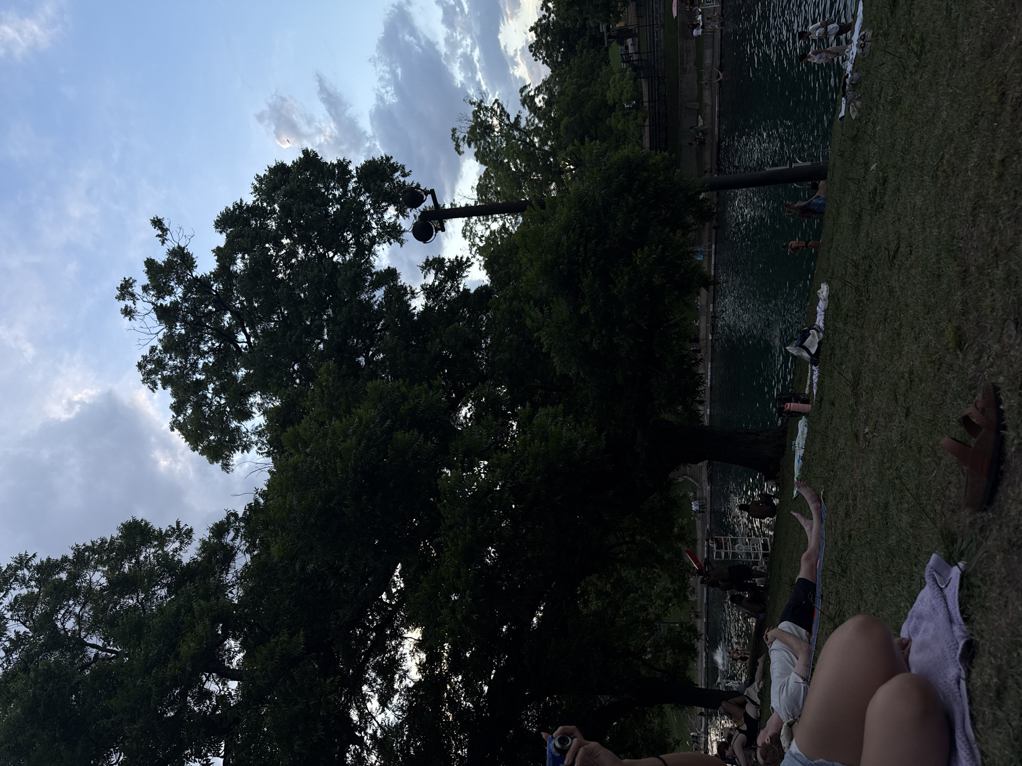 The image captures a scene at Barton Springs Pool in Austin, Texas. The pool is partially visible, bordered by grassy areas where people are relaxing. Some individuals are in the water, while others are lounging on towels on the grass. Trees surround the area, and a light pole is positioned among the trees. The sky is visible, with clouds scattered across it. A person's legs and parts of their clothing are visible in the foreground.