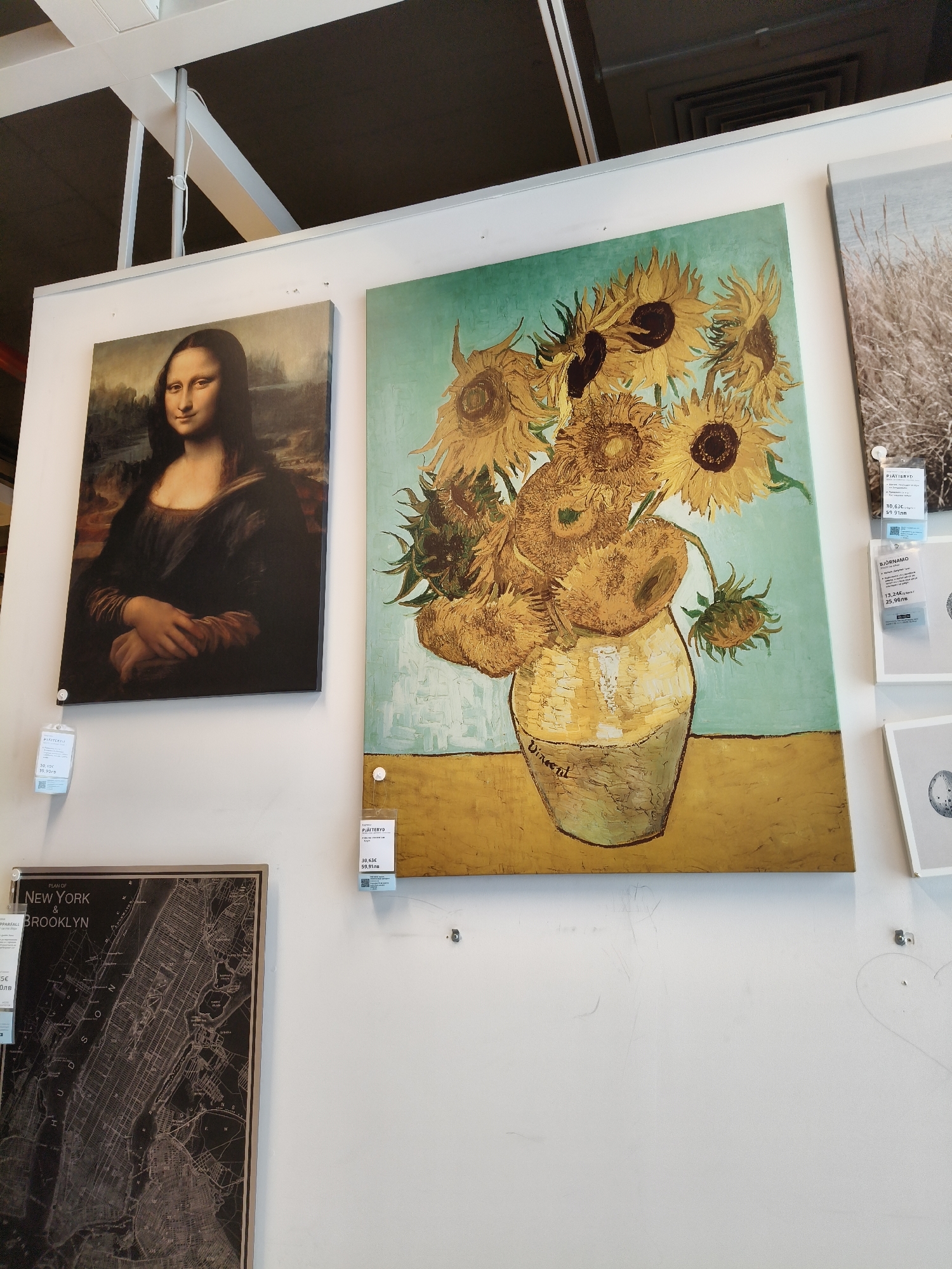 The image shows four framed pictures hanging on a white wall in a store. From left to right, the first picture is a reproduction of Leonardo da Vinci's Mona Lisa, with a price tag attached below. The second picture is a black-and-white map of New York and Brooklyn with a price tag attached. The third picture is a reproduction of Van Gogh's Sunflowers, also with a price tag attached. The fourth picture depicts dried grass in black and white, with a price tag below. There are also two smaller framed pictures to the right of the grass picture, with price tags attached. A drawing of a heart can be seen on the wall below the Sunflower painting.