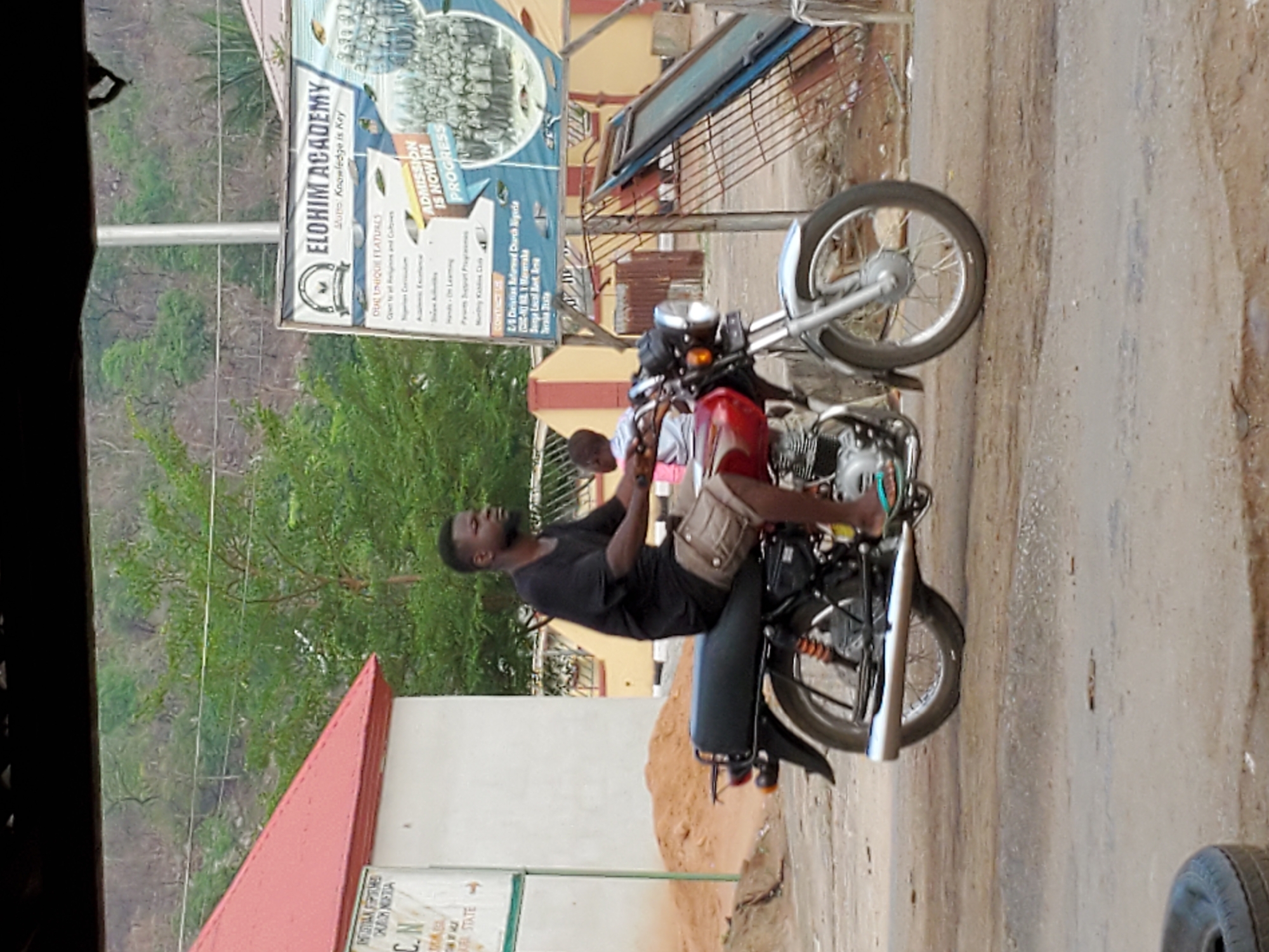 A man in a black t-shirt sits astride a parked red motorcycle on a dirt and paved road in Mararaba, Nigeria. He wears a tan satchel. A sign for Elohim Academy is visible in the background, advertising admission. Another sign is partially visible in the foreground. Trees and a hillside are behind the buildings.