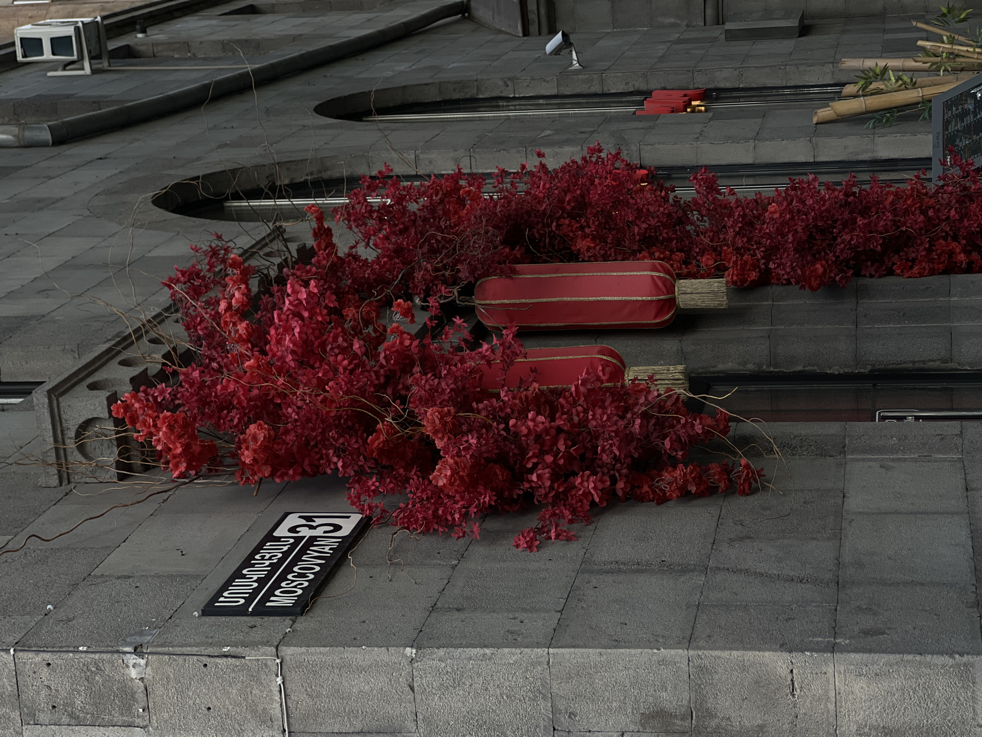 The image captures an outdoor architectural detail, possibly a multi-level facade or retaining wall, constructed from large, rectangular grey tiles. The predominant feature is a vibrant decorative arrangement of bright red dried or artificial foliage, scattered across different levels of the grey surface, providing a stark color contrast.
Interspersed within the red foliage are two red, cylindrical lanterns, each embellished with golden trim and featuring a bundled, light-brown wooden-stick design at its ends. These lanterns are placed horizontally among the red leaves.
In the lower-left section of the image, a black and white street sign is prominently displayed. It reads "31" and "MOSCOVYAN" in English uppercase letters, with the corresponding Armenian script written above it.
Other details include a bundle of bamboo stalks with some green leaves visible in the upper right quadrant. In the upper left, a white ventilation unit and a small security camera are mounted on the grey structure. The lighting is even, suggesting an overcast day or a shaded area. No people or active events are depicted; the scene is a static decorative display. Given the visible text and provided context, the location is Moskovyan Street 31 in Yerevan, Armenia.
