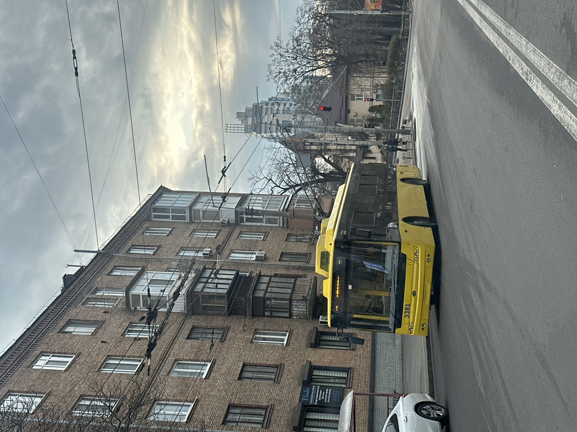 The image depicts a street scene in Kyiv, Ukraine. A yellow trolleybus, numbered 3381, is parked on the right side of a paved road, adjacent to a brick building with multiple windows and balconies. Electrical wires for the trolleybus system run above the vehicle and along the building. A red traffic light is visible in the background, near another structure with a visible roof. The sky is overcast. A white car is partially visible parked behind the trolleybus.