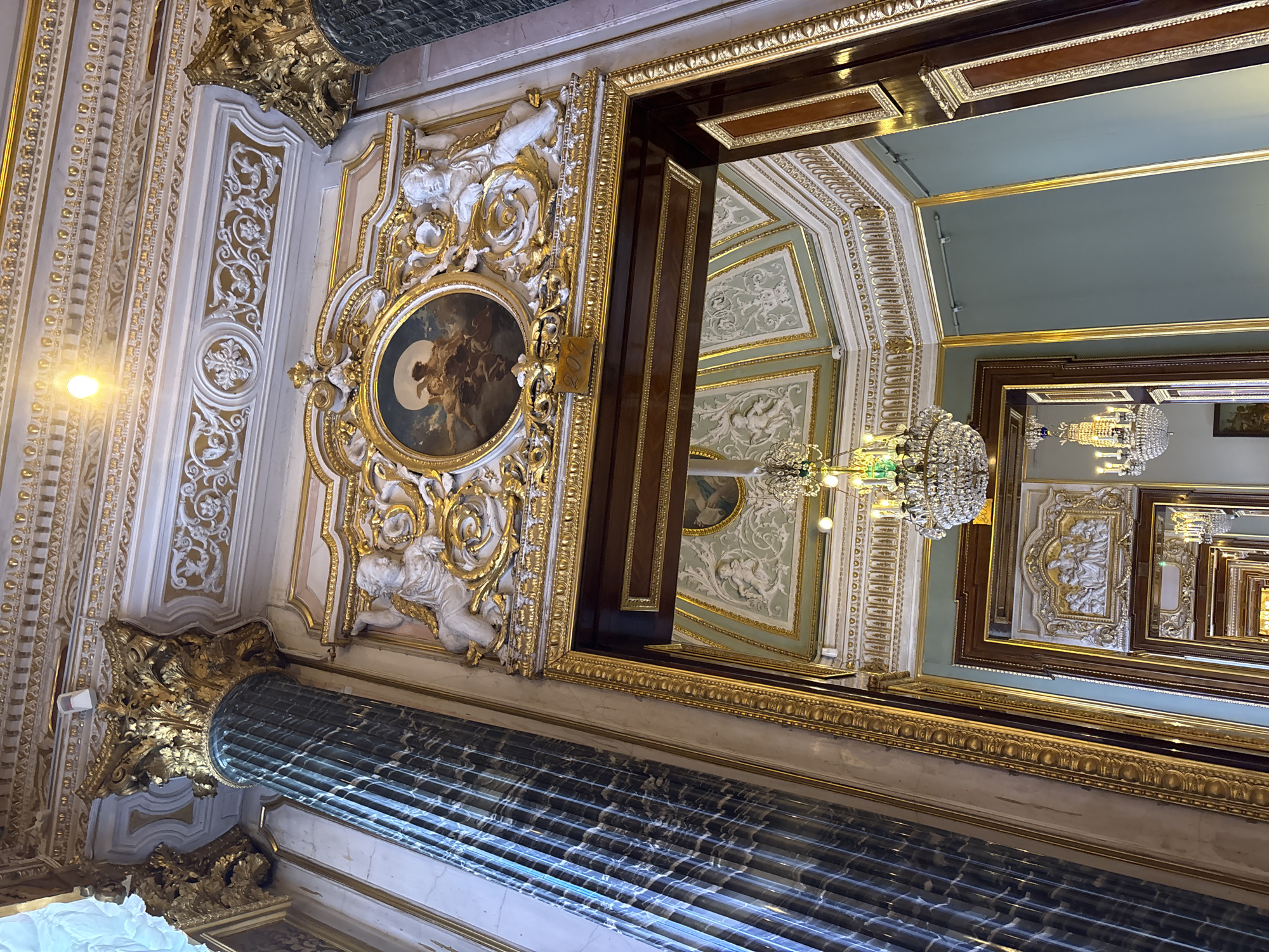The image depicts the opulent interior of a grand hall, likely in the Hermitage Museum in Saint Petersburg, Russia. The scene is characterized by rich ornamentation, including ornate gold detailing, detailed white stucco moldings, and a large, green marble column with a golden Corinthian capital on the left. Above a doorway, a circular painting is set within an elaborate golden frame adorned with cherubic figures and scrolling acanthus leaves. The ceiling is intricately decorated with plasterwork and gold accents, and a crystal chandelier hangs prominently. Beyond the doorway, another hall is visible, featuring mirrored walls and more chandeliers, creating a sense of depth and grandeur, with a repeating pattern of decorative moldings and recessed panels. The color palette is dominated by gold, white, and a pale green for the walls, contributing to an atmosphere of historical luxury.