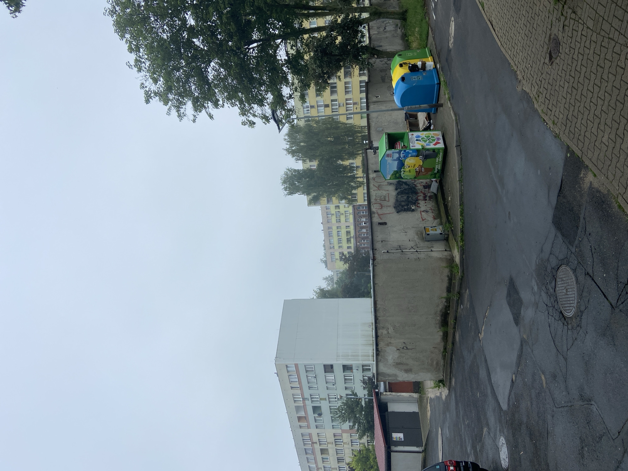 This is a daytime, eye-level, medium shot of an overcast, outdoor scene in Bolesławiec, Poland. The image depicts a concrete wall in the foreground, with graffiti visible on its surface. To the right of the wall are three brightly colored recycling bins: one green, one blue, and one yellow. The green bin has a cartoon graphic depicting animated characters. Behind the wall, there is a paved area with manhole covers. Further in the background, a line of trees separates the recycling bins from a multi-story apartment building with light-colored walls and numerous windows. To the left of this building is a smaller, lighter-colored structure with a red roof, resembling a garage or storage unit. The sky is a uniform light gray, suggesting cloudy or foggy weather. The overall scene is one of an urban residential area, likely a courtyard or communal space.