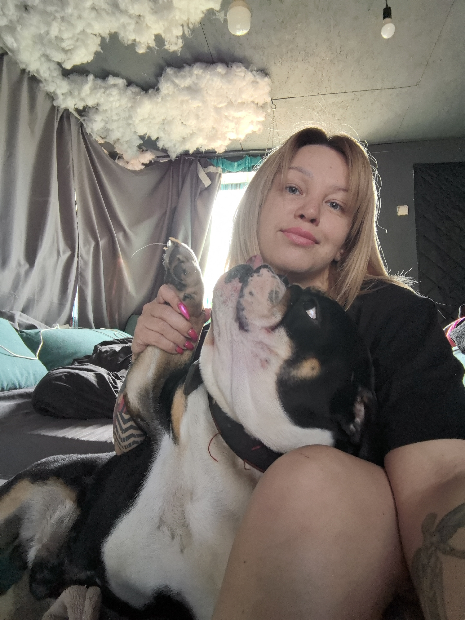 This indoor photograph captures a heartwarming moment between a young woman and her dog. The woman, with shoulder-length blonde hair, fair skin, and light blue eyes, gazes calmly at the camera with a subtle, content smile. She is dressed in a dark top, and her right hand, featuring bright pink nail polish, gently holds the front paw of her pet.
Beside her, a dog, likely a bulldog or a similar mixed breed with black, white, and tan markings, looks up at the woman with an affectionate expression, its mouth slightly open as if licking its nose. They are positioned closely, suggesting they are relaxing together on a bed or sofa, indicated by the teal and dark-colored pillows visible in the background.
The room is illuminated by bright natural light, implying it is daytime. Grey curtains are drawn across a window area, while the ceiling above is uniquely decorated with fluffy, white cloud-like material, creating an ethereal ambiance. Two simple light bulbs also hang from the ceiling. The woman has visible tattoos on her arm and leg. The overall scene conveys a sense of comfort and affection shared between the woman and her canine companion within a distinctively decorated home in Noviy, Russia. No text is visible in the image.