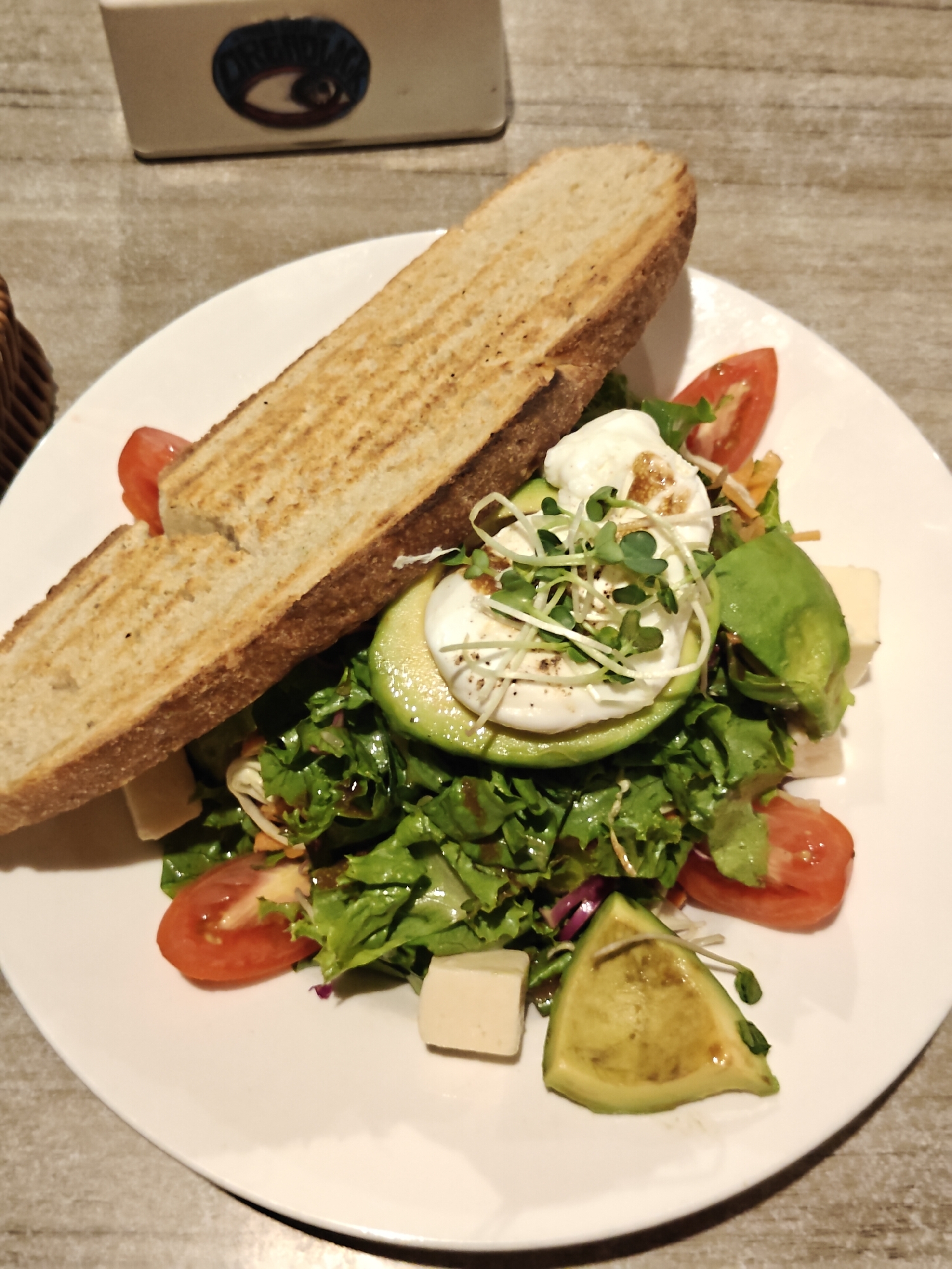 A close-up, high-angle shot captures a white plate of food presented on a light-colored, textured surface, likely a table. The dish features a salad with mixed greens, cherry tomato halves, cubed white cheese, and sliced avocado. Atop the salad is a poached egg, garnished with microgreens and a drizzle of dressing. Two slices of toasted bread are arranged diagonally across the plate. In the background, a portion of a beige, rectangular object with a circular emblem is visible. The overall presentation suggests a meal served in a casual dining setting. The location is identified as Pokhara, Nepal, implying this culinary scene is set within that city.