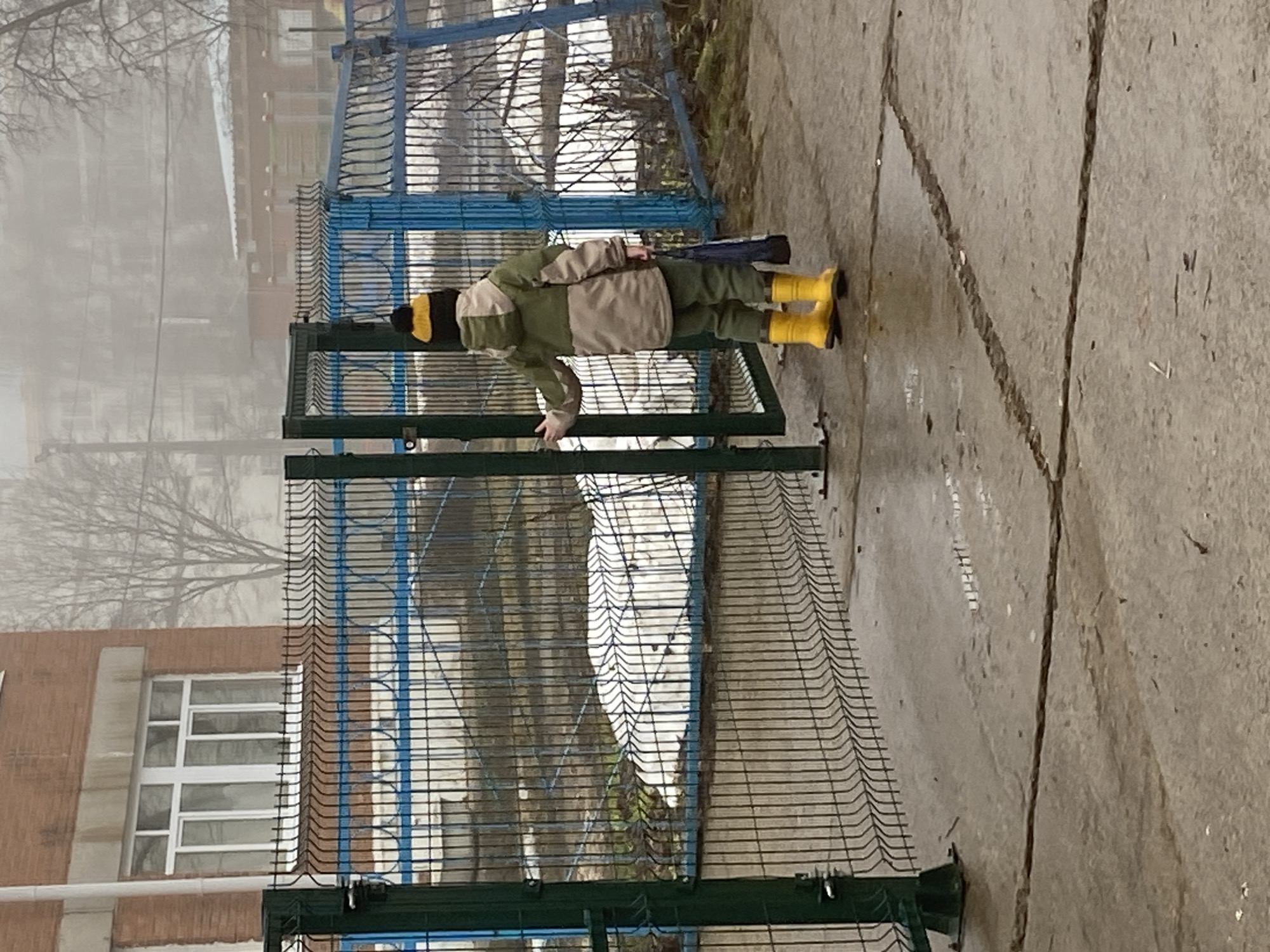 A child is standing next to a blue and green metal fence on a concrete surface. The child is wearing a green and beige jacket, green pants, a black and yellow hat, and yellow boots. The fence appears to surround a building with windows. In the background, there is a building and trees obscured by fog. The scene appears to be in an urban environment, potentially Noviy, Russia.